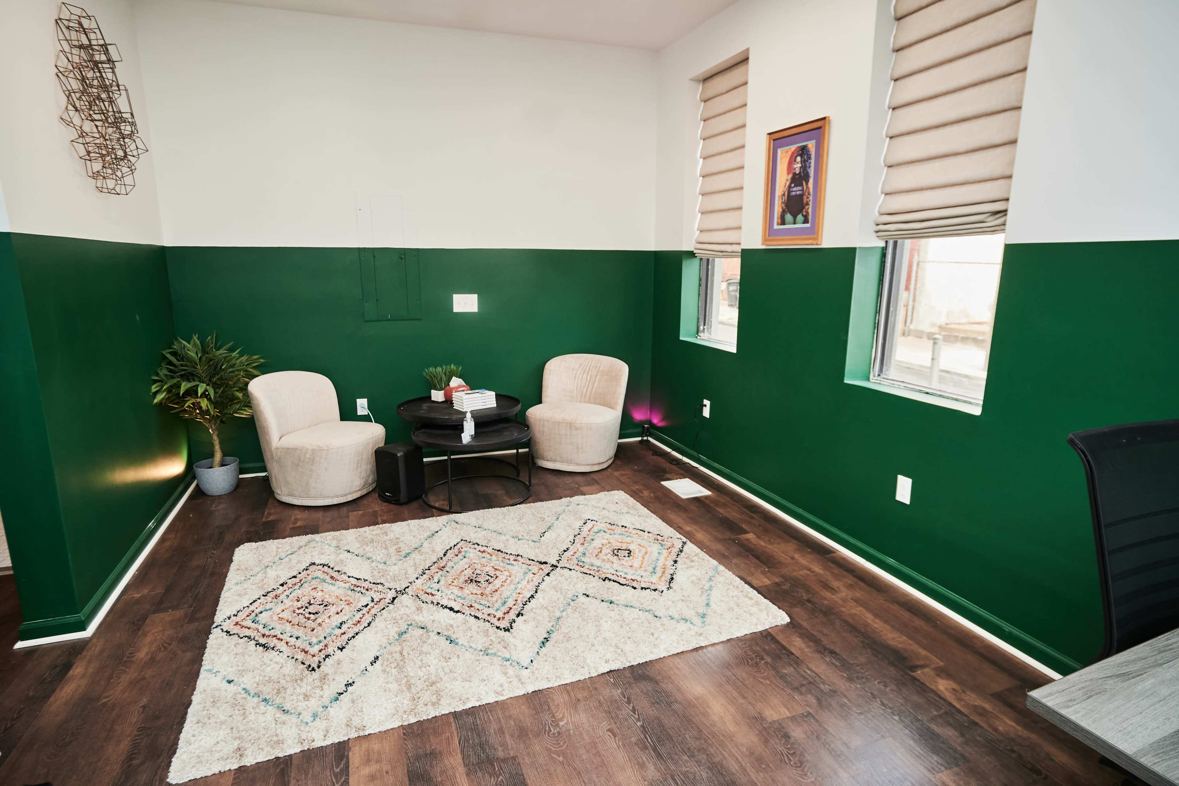 A room with green and white walls, two beige chairs, a small black table, a patterned rug, and windows with blinds.
