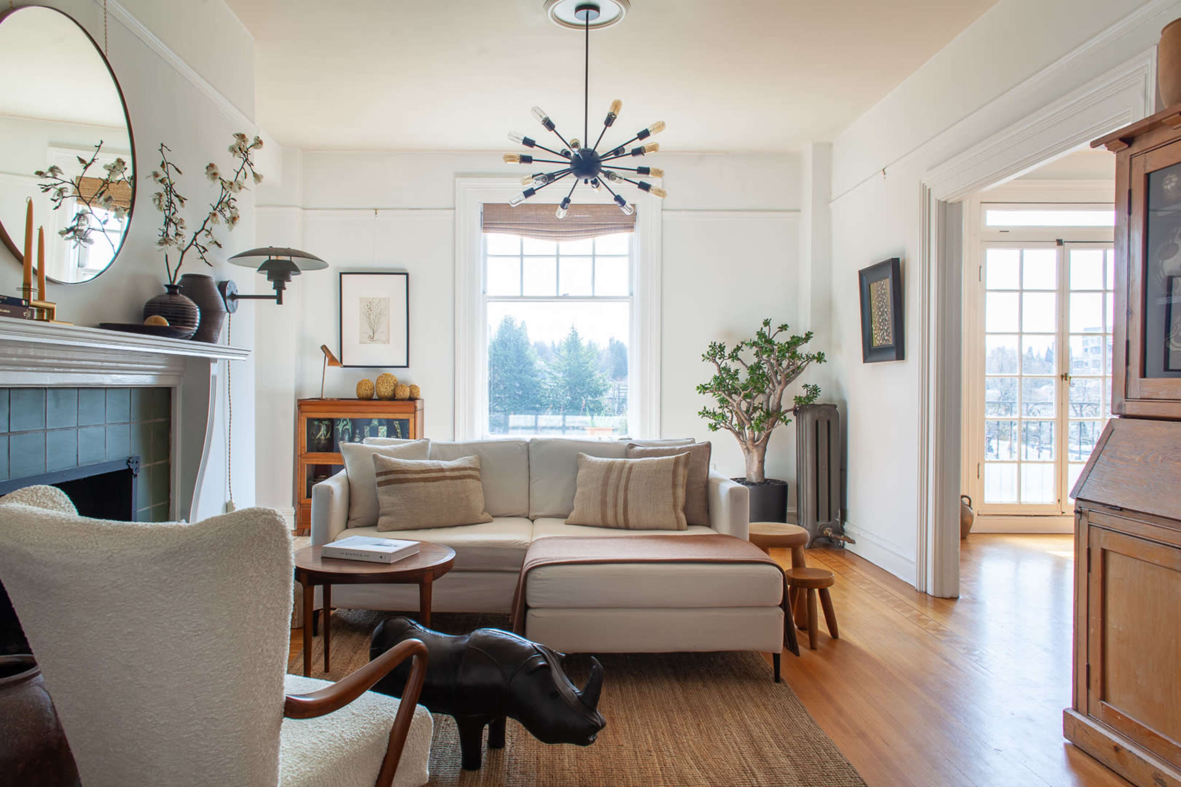 The image shows a bright living room featuring a modern sofa, an armchair, and large windows with a view of trees outside.