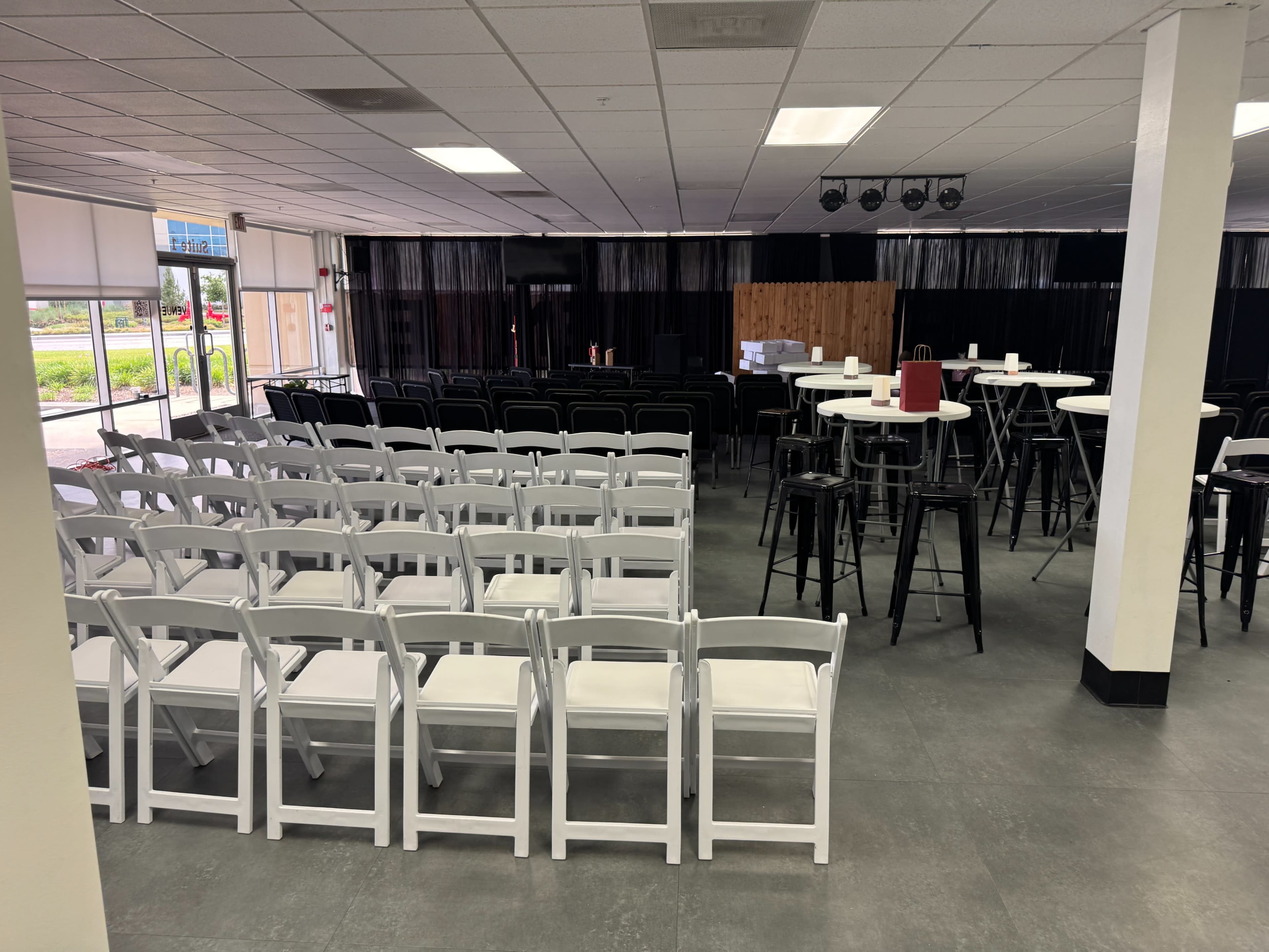 The image shows a spacious event venue with rows of white folding chairs, high-top tables, and a black curtain backdrop.