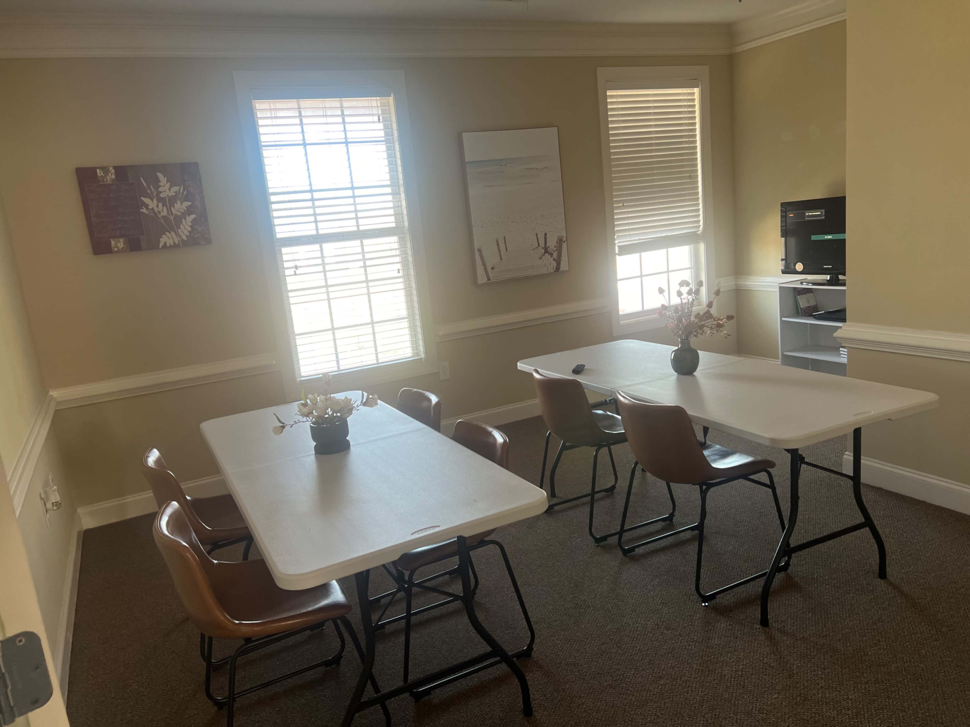 The image shows a small meeting room with two foldable tables and several chairs, featuring a flower centerpiece and a large window with blinds.