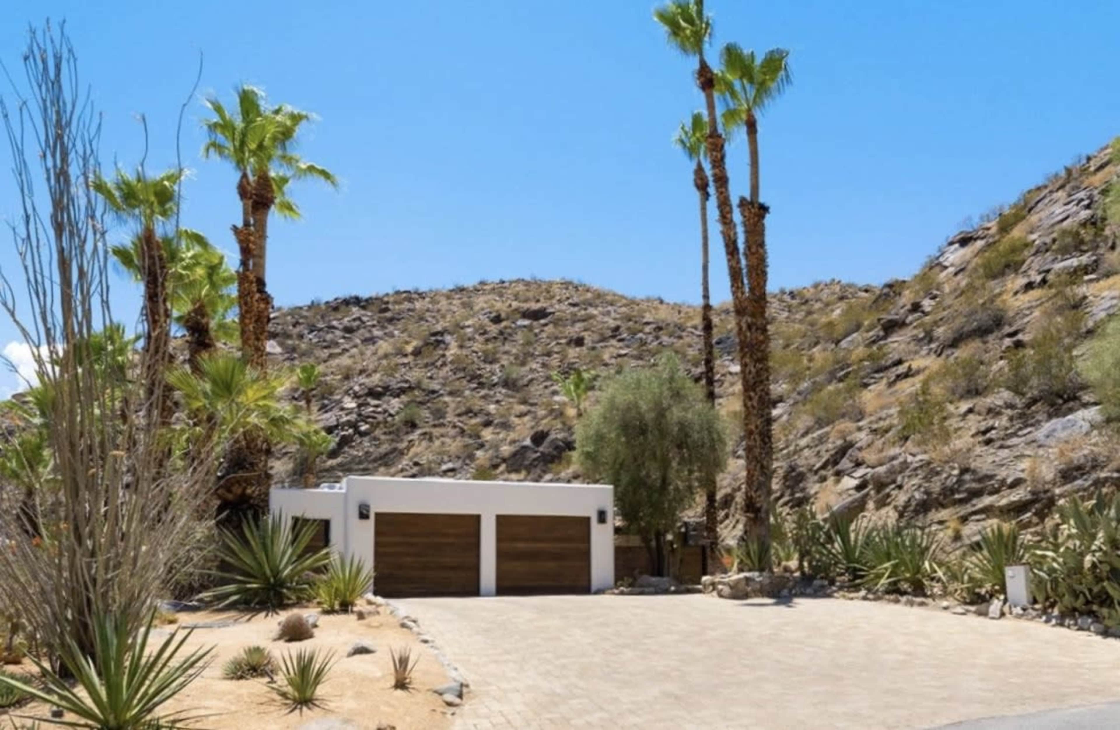 The image shows a modern home with a garage, surrounded by desert landscaping and palm trees, set against a rocky hillside.