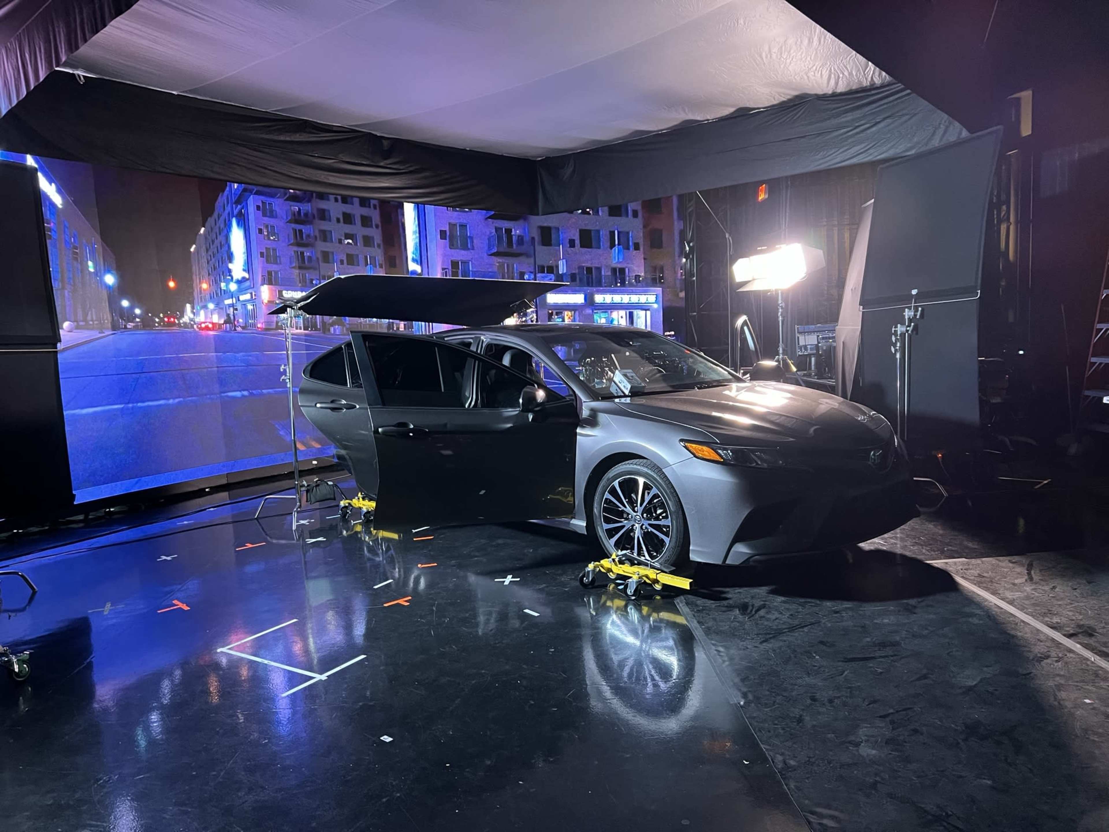 A black sedan is positioned on a stage with artificial lighting and a large digital backdrop depicting a city street at night.