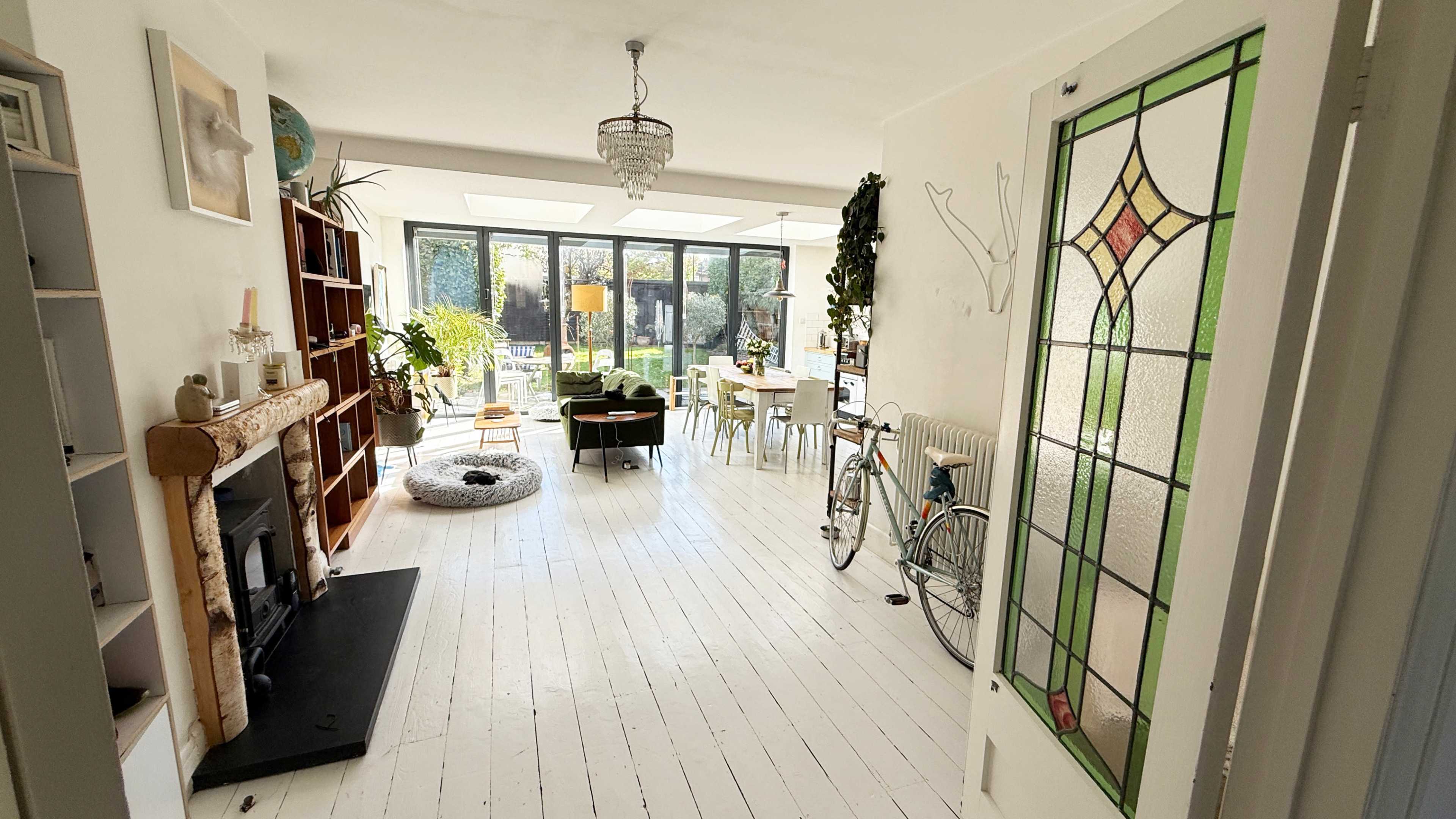 The image shows a spacious, well-lit living room with white wooden floors, a fireplace, a bicycle, and a dining area surrounded by plants and large windows.
