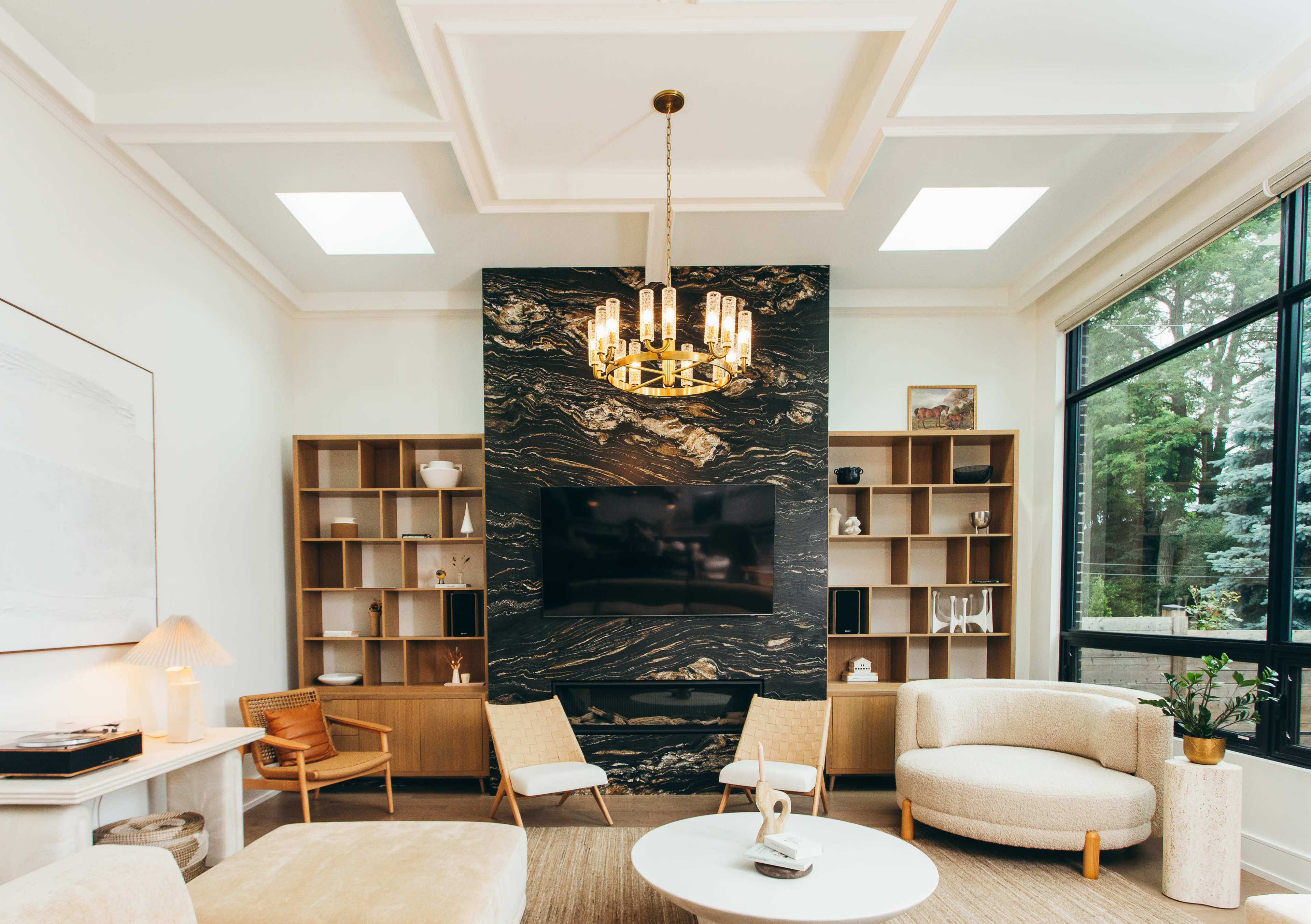 A modern living room features a decorative wall with a large TV, surrounded by built-in shelves, a round coffee table, and two chairs, illuminated by a stylish chandelier.