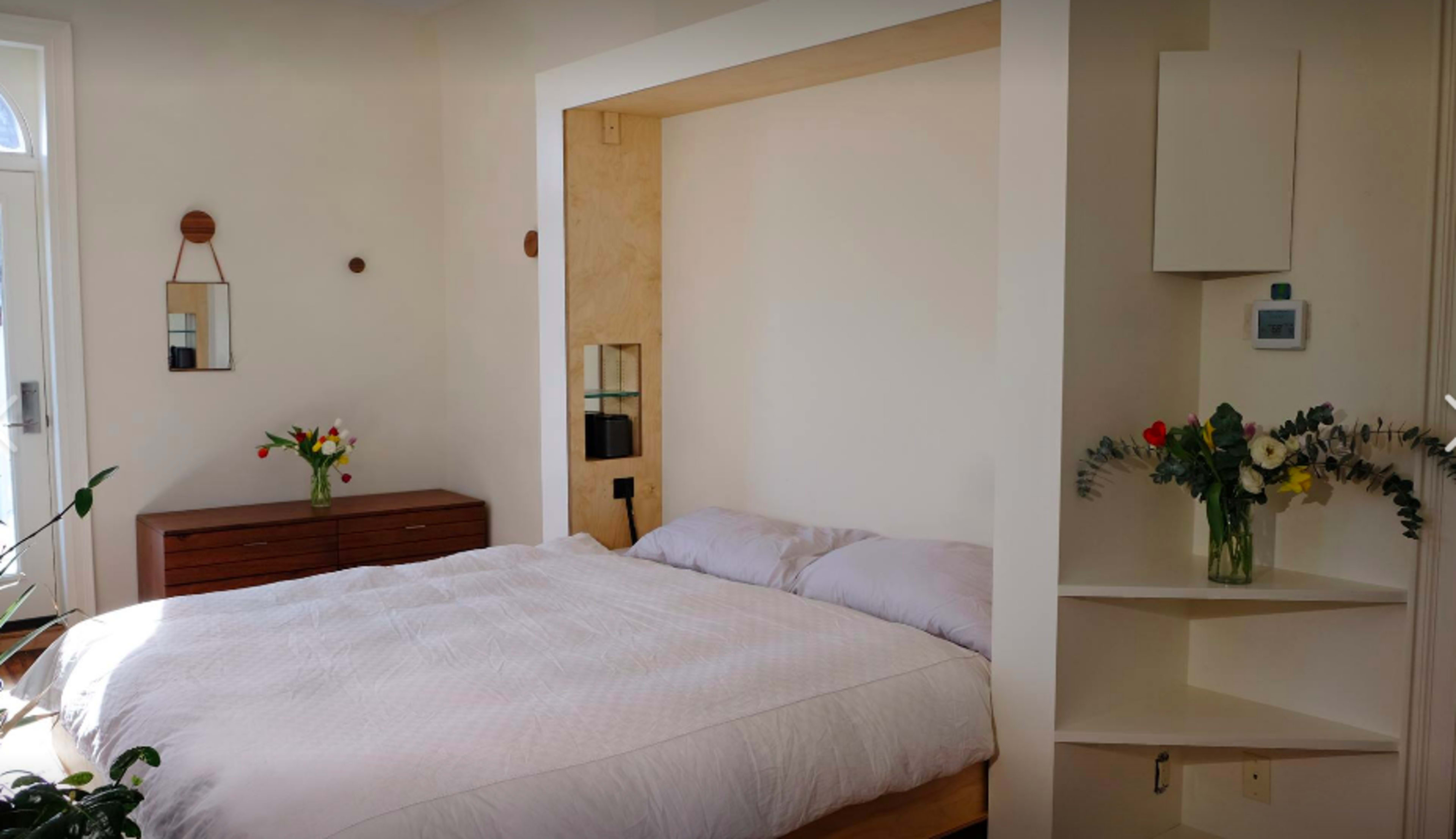 A minimalist bedroom featuring a bed with light bedding, a wooden dresser, and decorative plants.