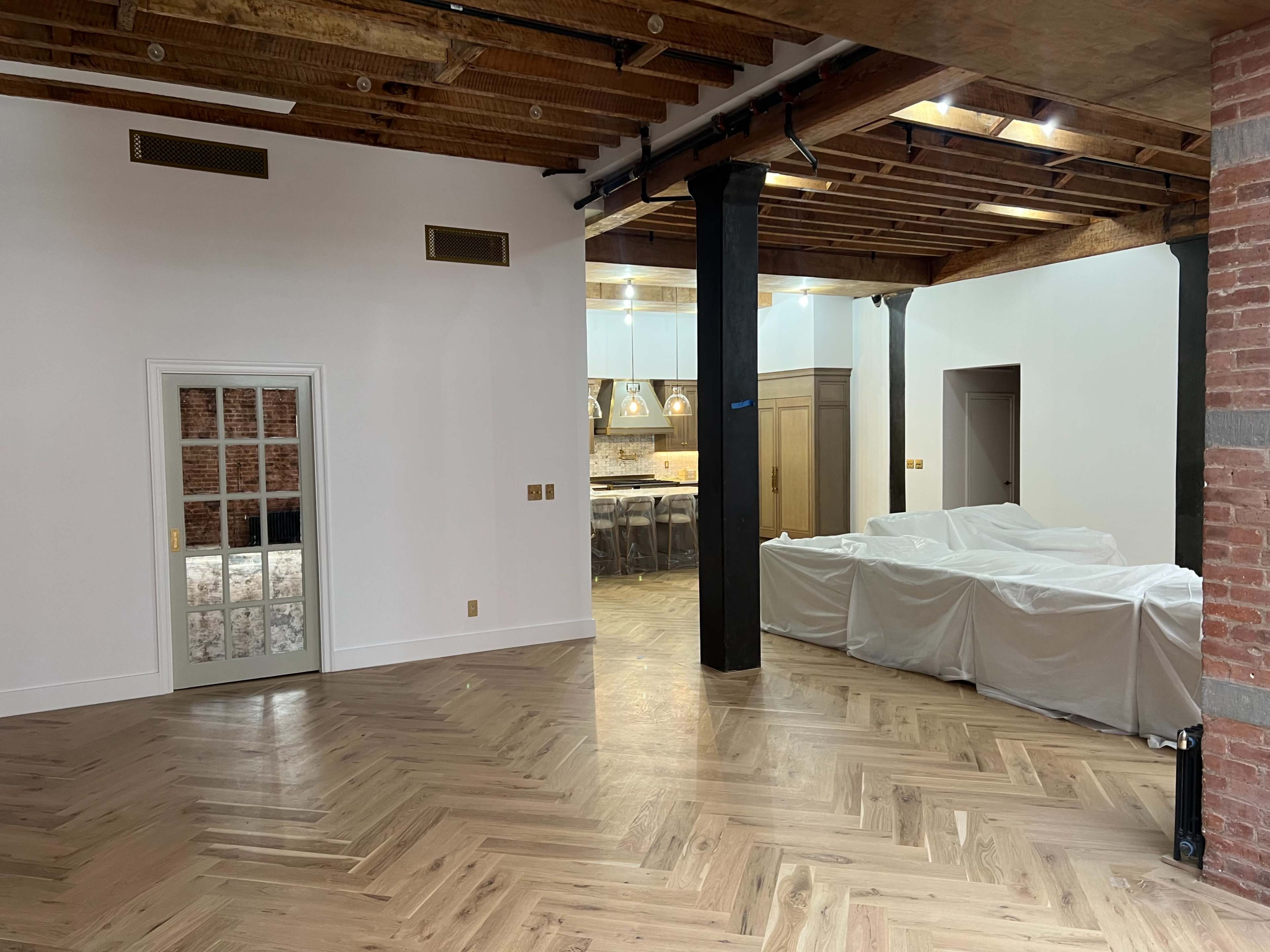 The image shows a spacious interior with wooden floors, exposed beams, a closed door with a glass panel, and a covered area on the right side.