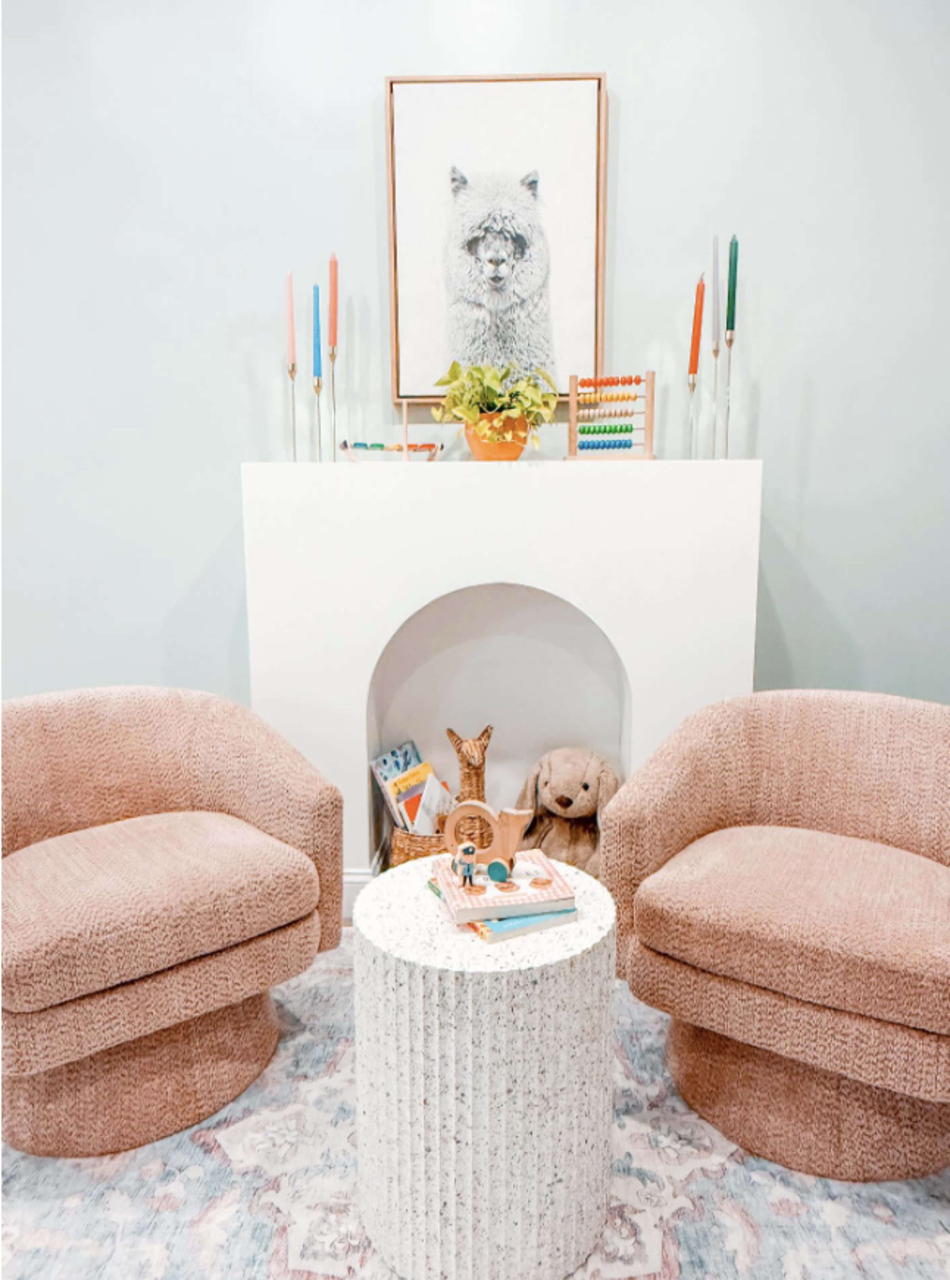 The image features a cozy seating area with two pink, rounded armchairs, a white pedestal table, and a wall-mounted artwork of an animal above a decorative mantle.