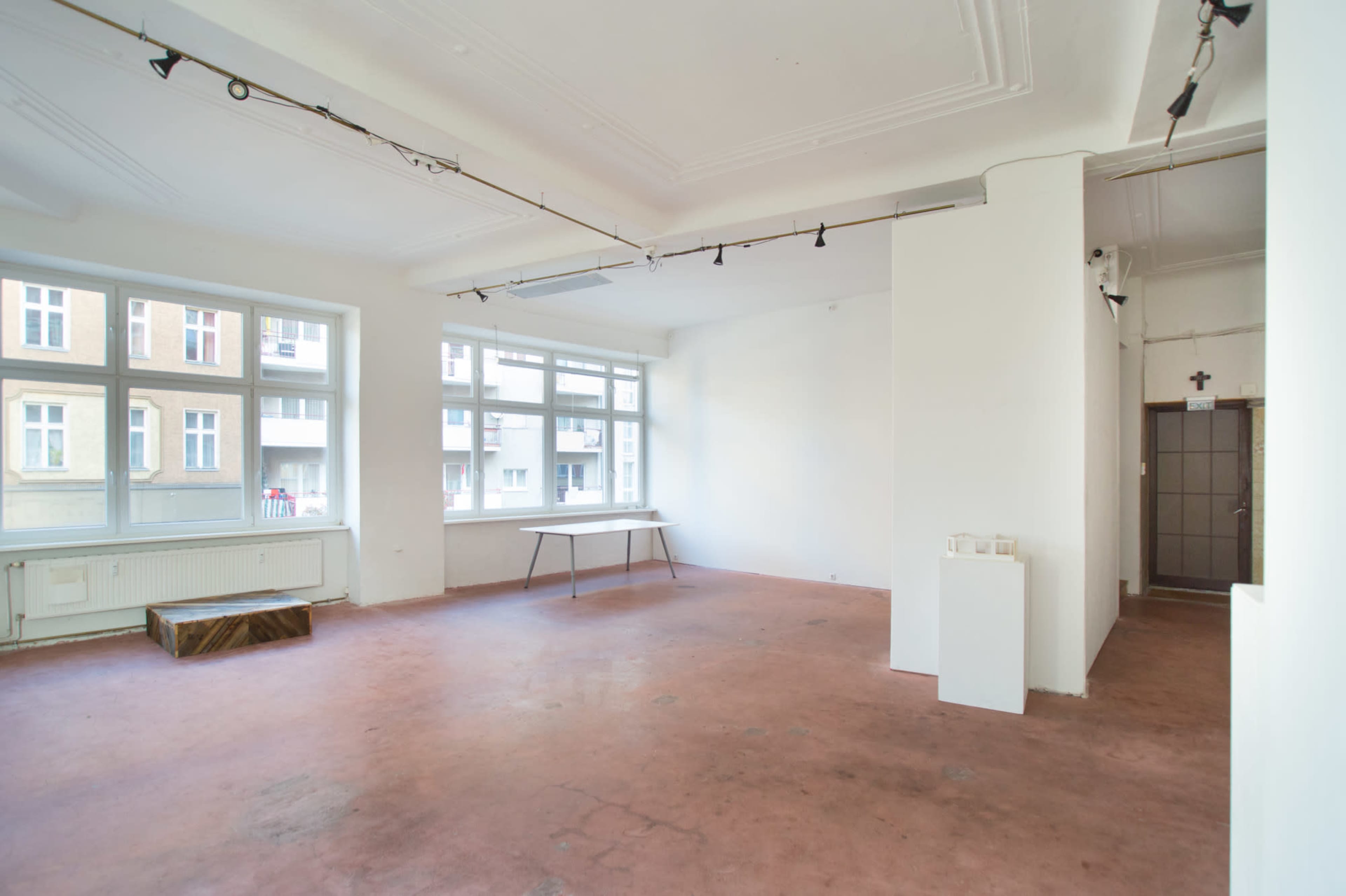 An empty room with large windows, a table in the center, and a white pedestal near the entrance.