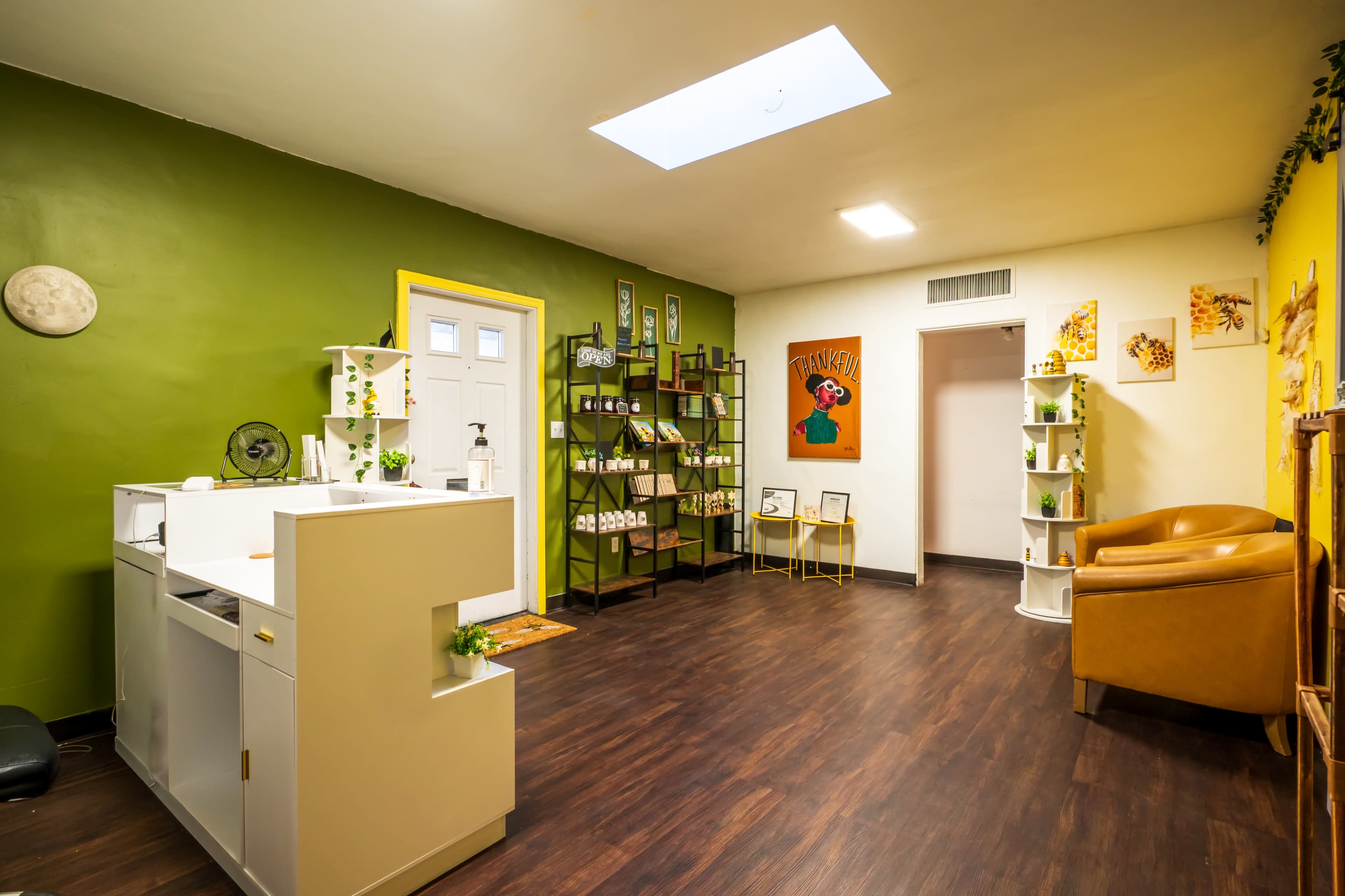 The image shows a well-organized room with green and yellow walls, featuring a reception area with a desk, shelves displaying products, a cozy seating area, and a skylight.