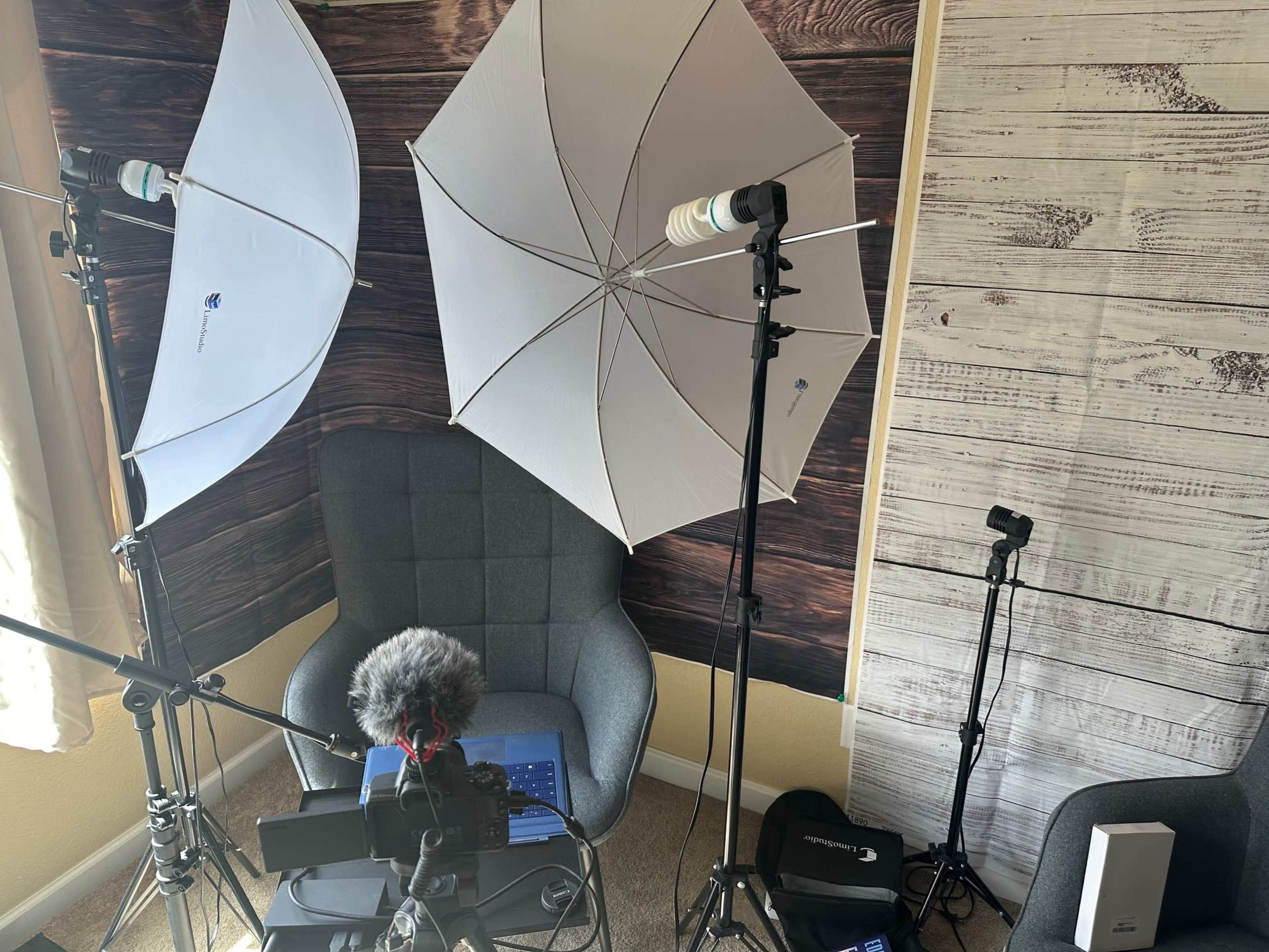 A gray chair surrounded by two umbrella lights, a microphone, a laptop, and a backdrop of wooden panels.