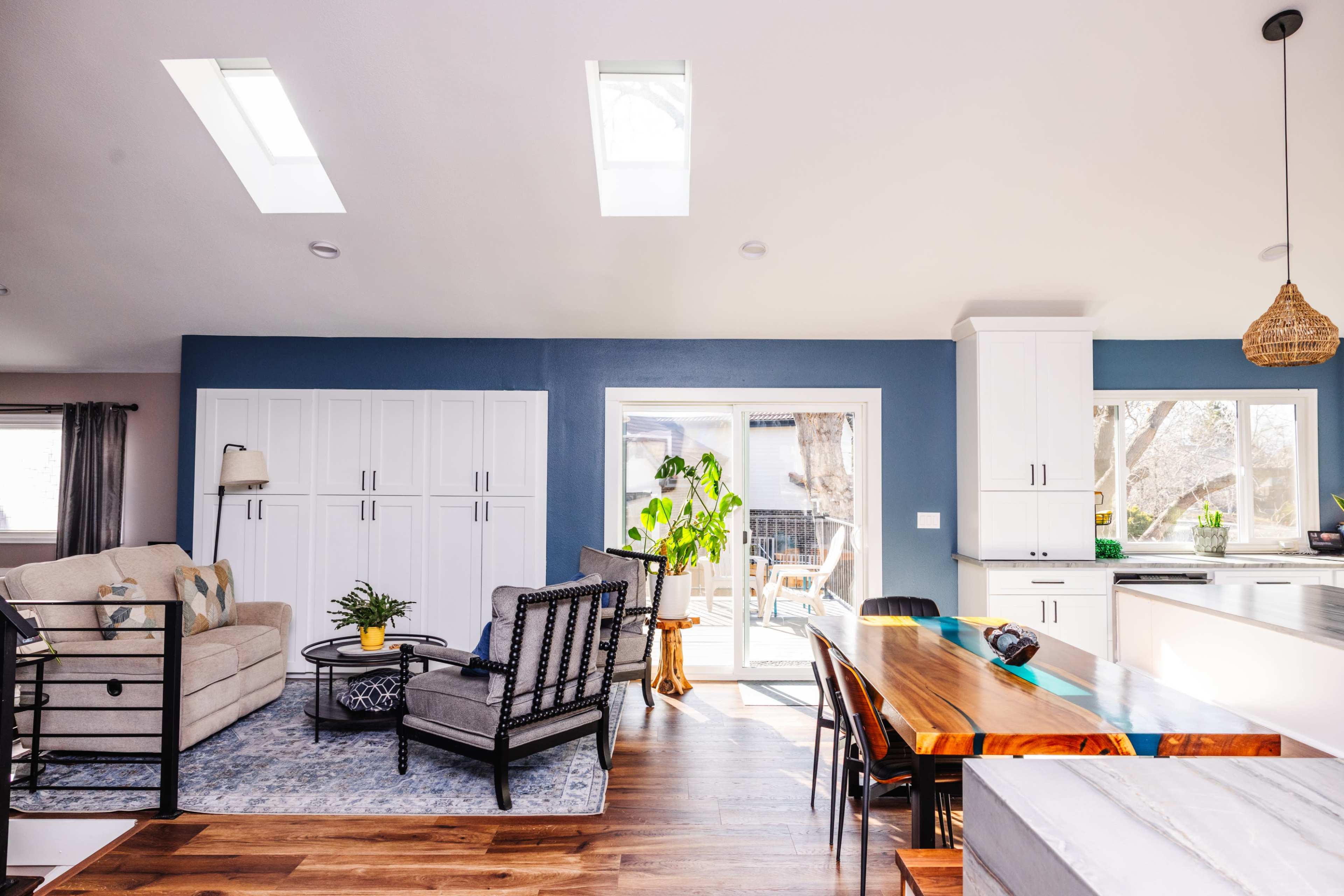 The room features a modern living space with a comfortable seating area, natural light from skylights, and a dining table made of wood, set next to sliding glass doors that open to an outdoor deck.