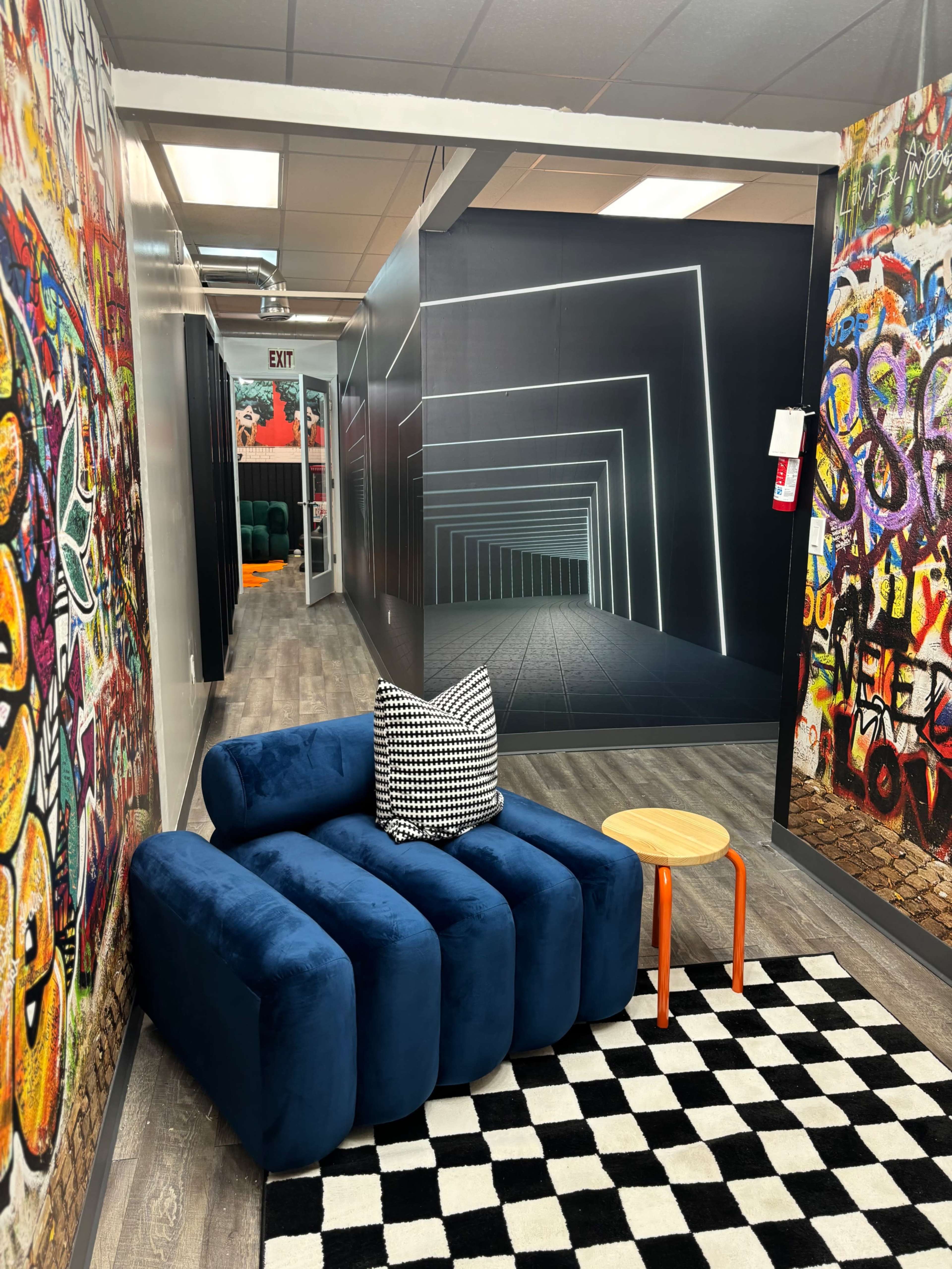 The image shows a modern lounge area featuring a blue, plush sofa with a patterned pillow and a small wooden table, surrounded by vibrant graffiti-style murals on one wall and a sleek, illuminated corridor in the background.