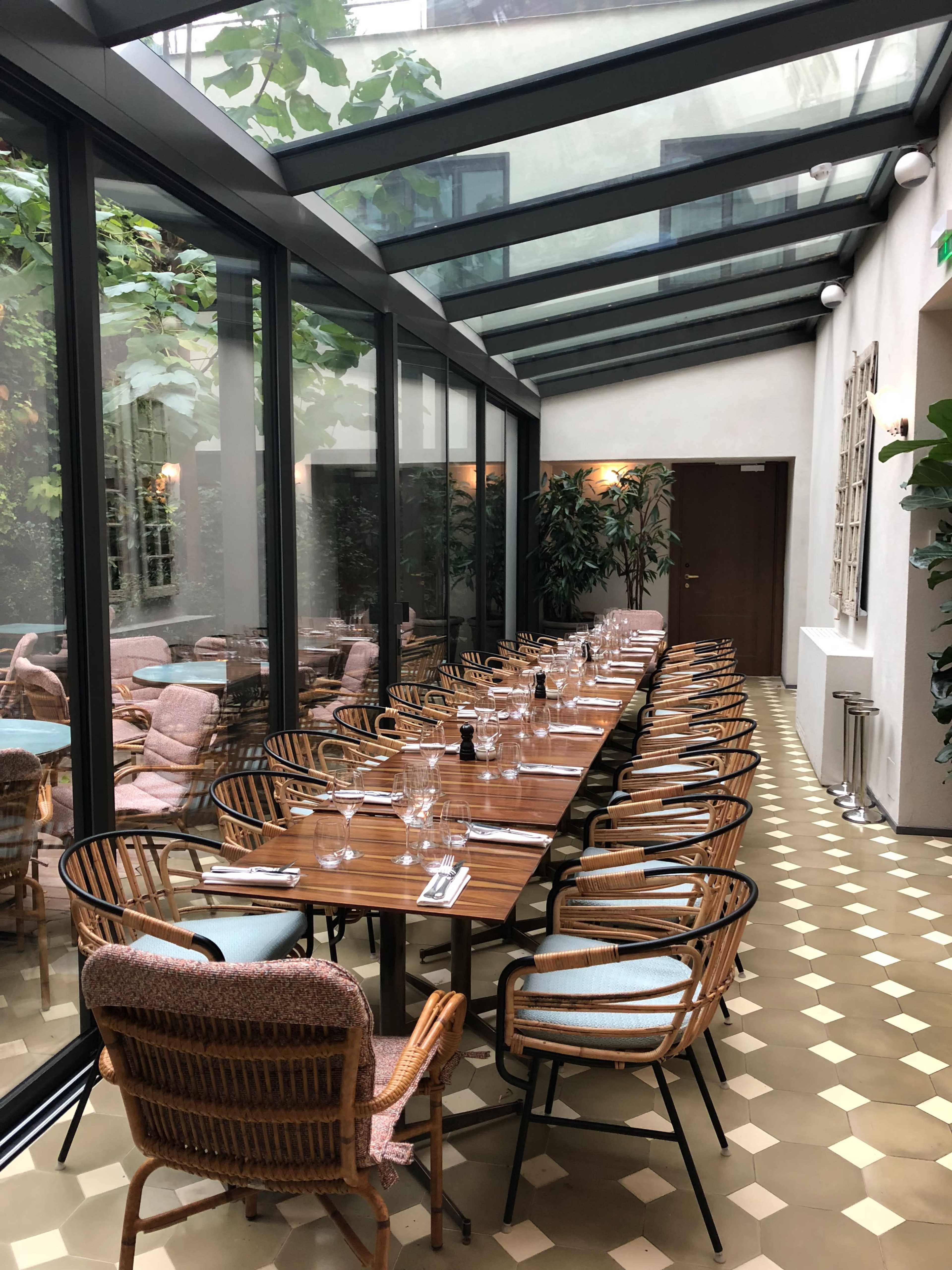 The image shows a dining area with a long, rectangular table set for a meal, surrounded by windows and potted plants.
