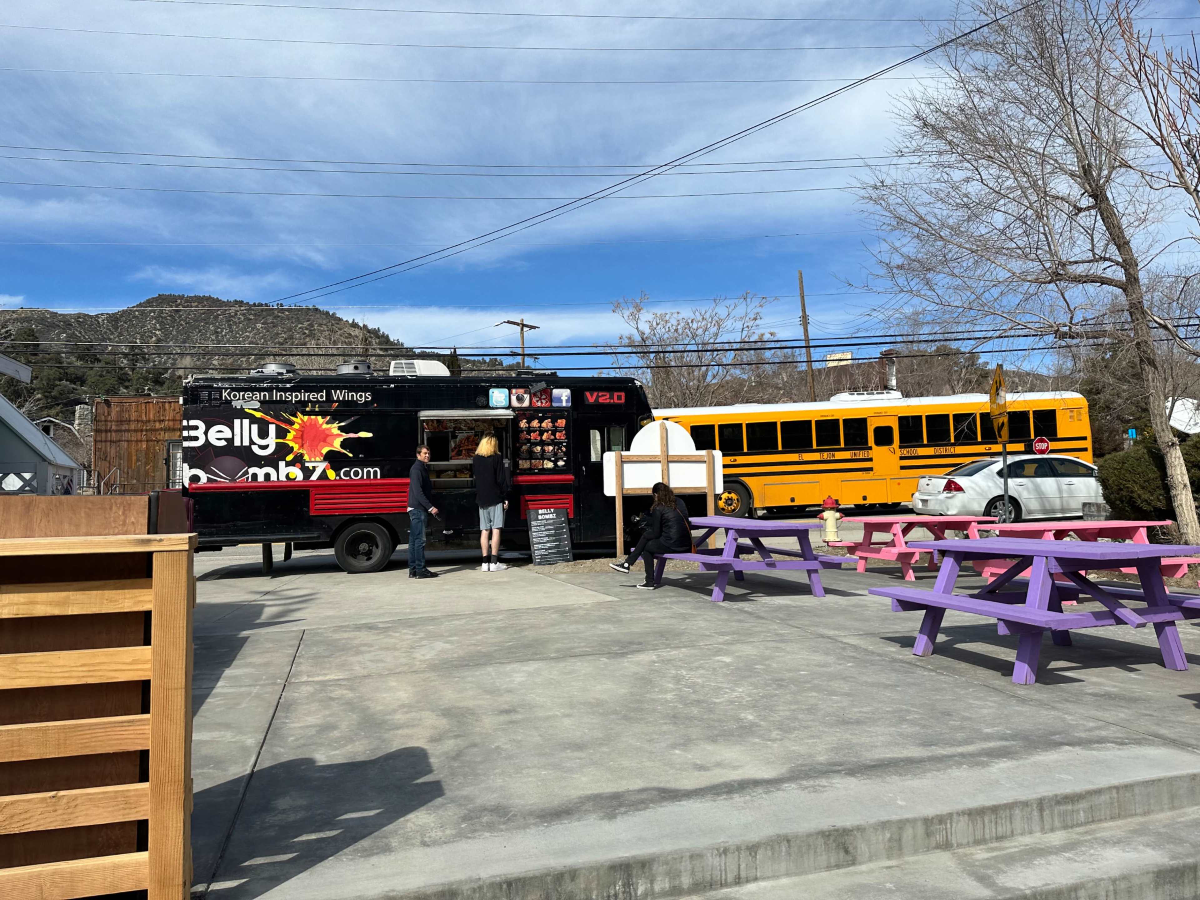 A food truck named "Bellybomb," offering Korean-inspired wings, is parked in a lot with several individuals ordering food, while a yellow school bus is visible nearby.