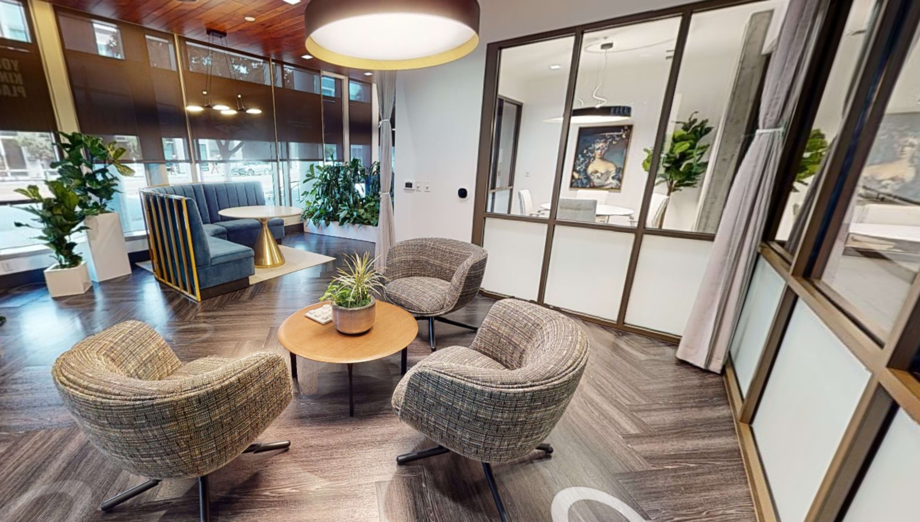 A modern office reception area with two pairs of chairs around a round table, a sofa in the background, and large windows featuring indoor plants.