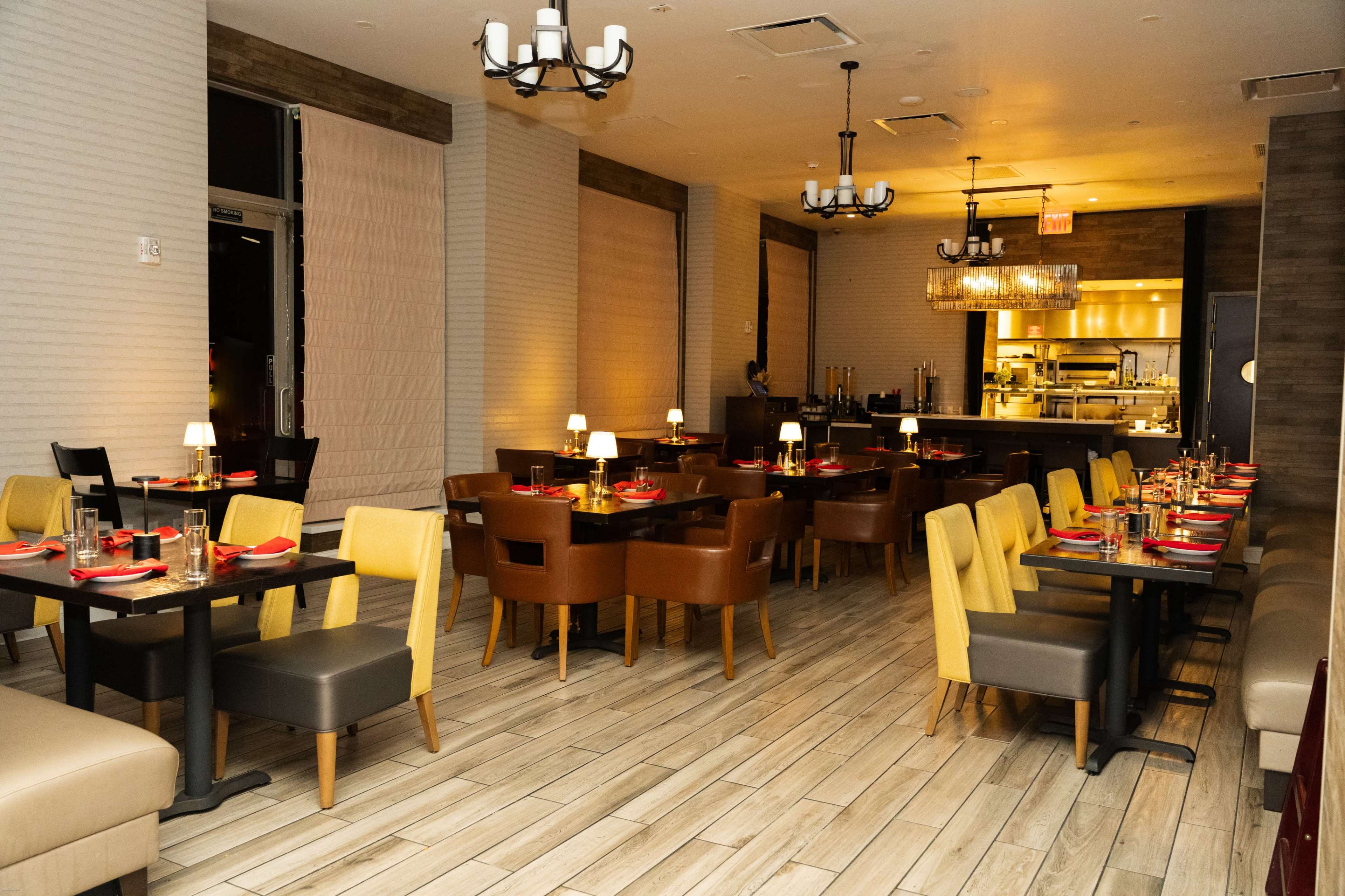 The image shows a modern restaurant interior with tables set for dining, featuring a mix of brown and yellow chairs, soft lighting, and an open kitchen area visible in the background.