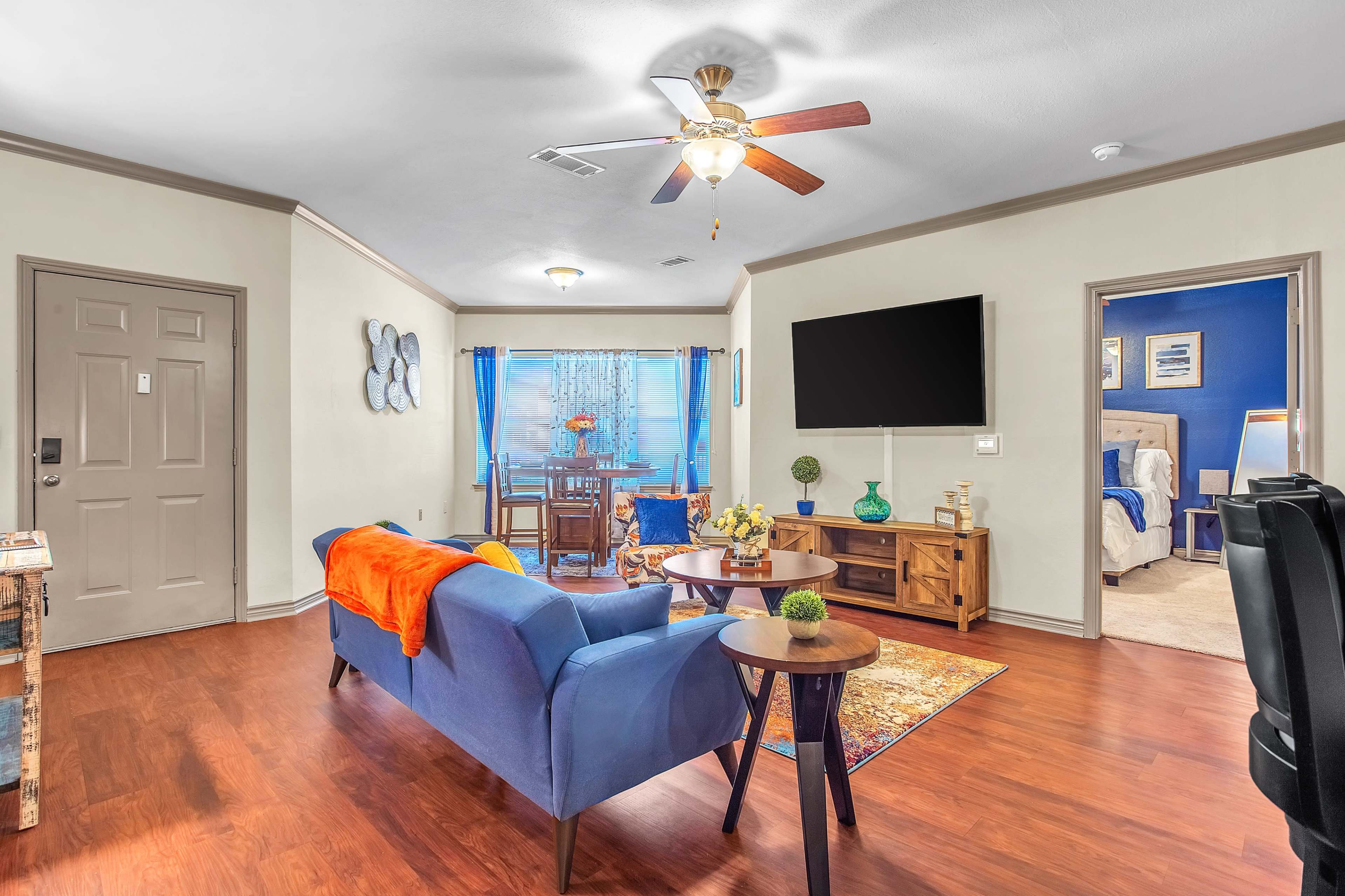 The living room features a blue sofa, a wooden coffee table, and a ceiling fan, with a view into a dining area and a bedroom through adjacent doors.
