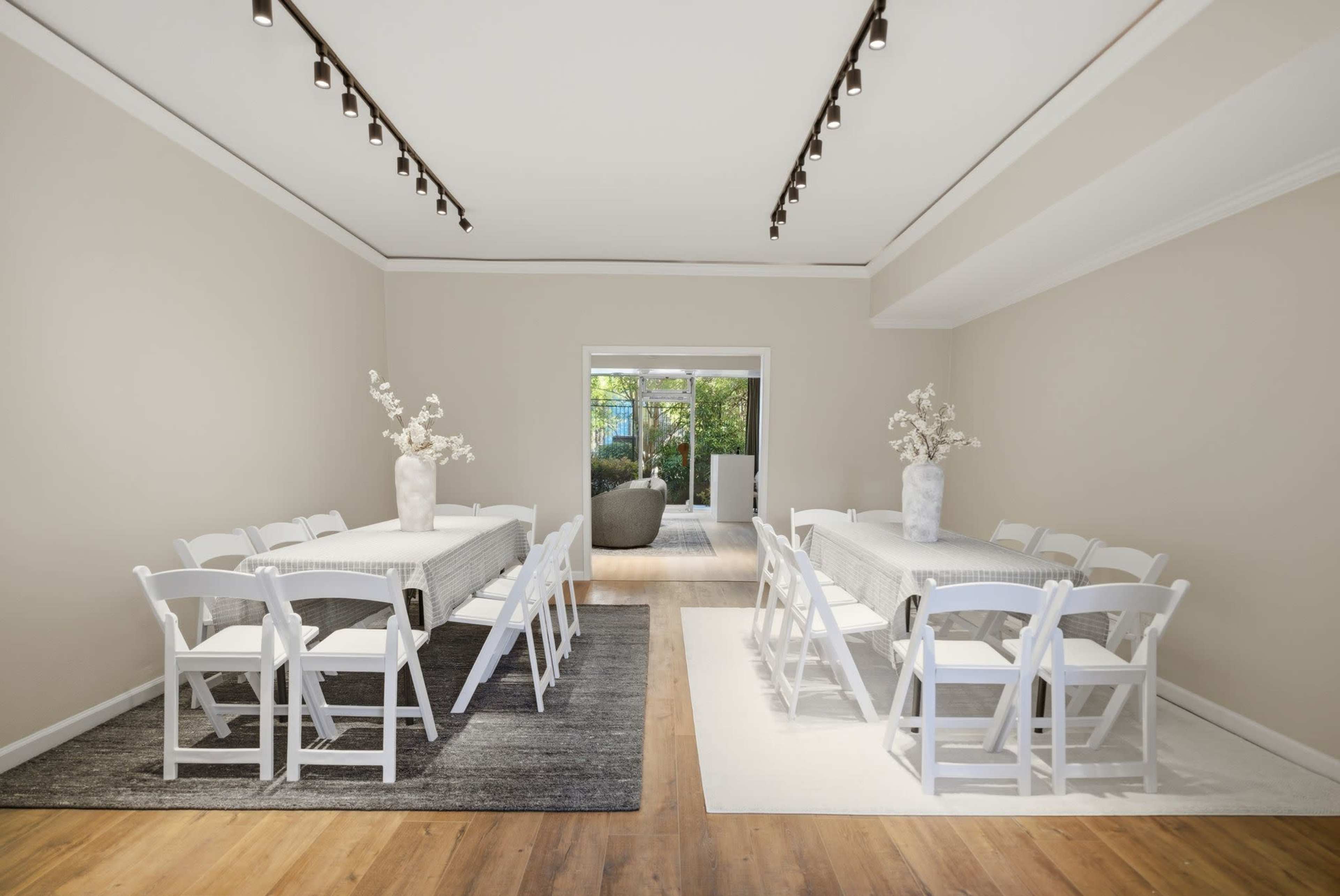 The image shows a spacious meeting room with two long tables and white chairs arranged on light-colored rugs, facing a glass door that leads to an outdoor area.