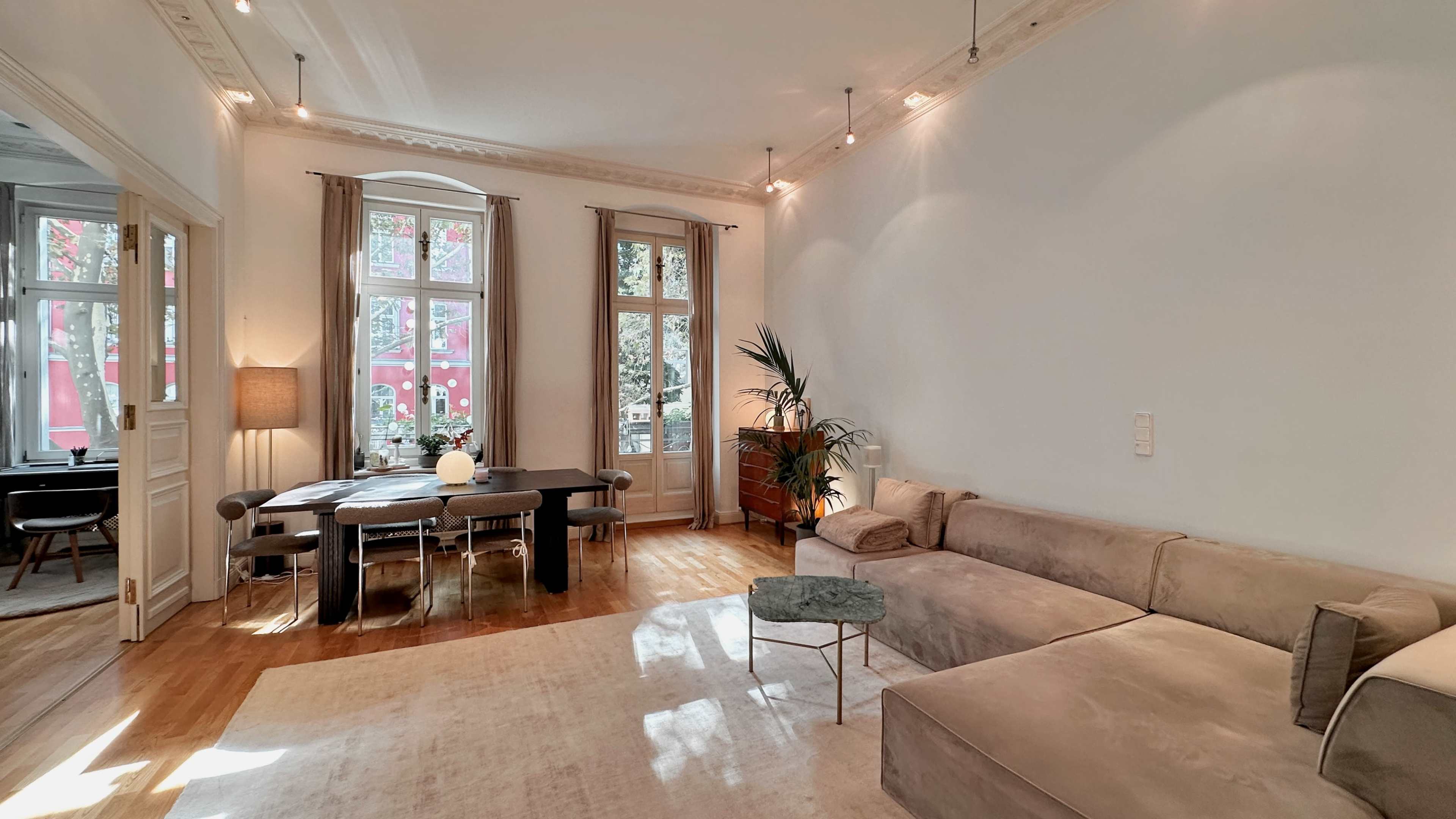 The image shows a bright, minimalist living room with a large sectional sofa, a small coffee table, and a dining area featuring a table and chairs near a window.
