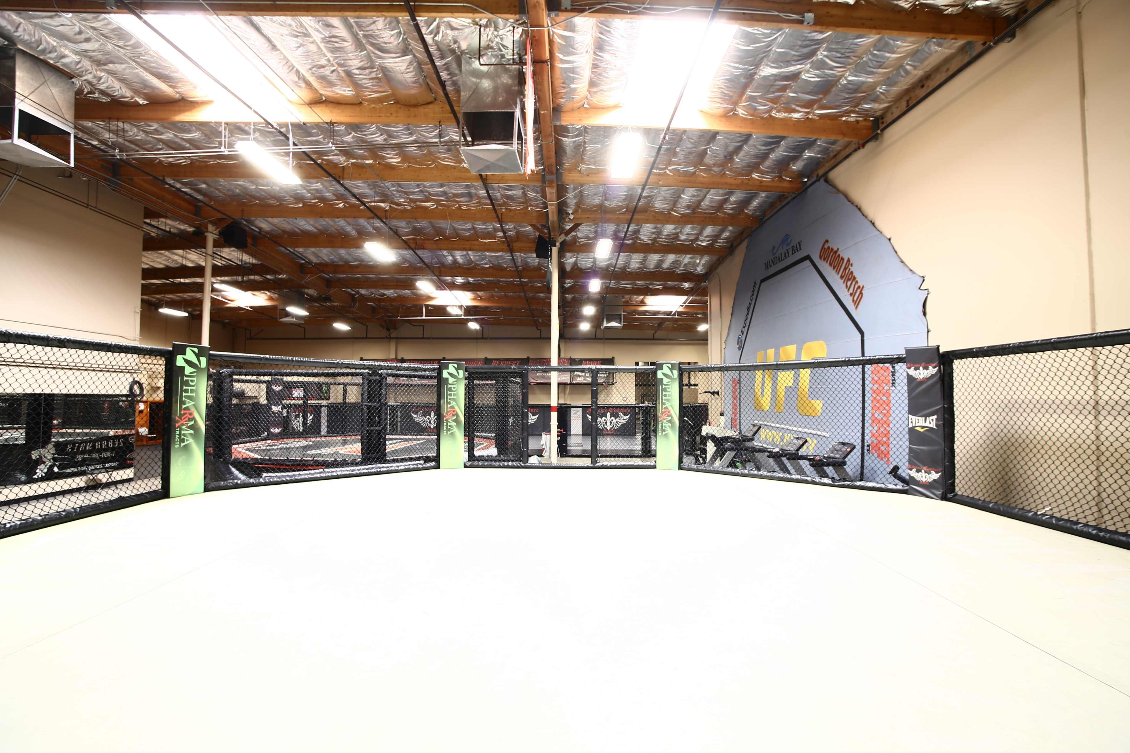 An empty training facility with a central octagonal cage and gym equipment in the background.