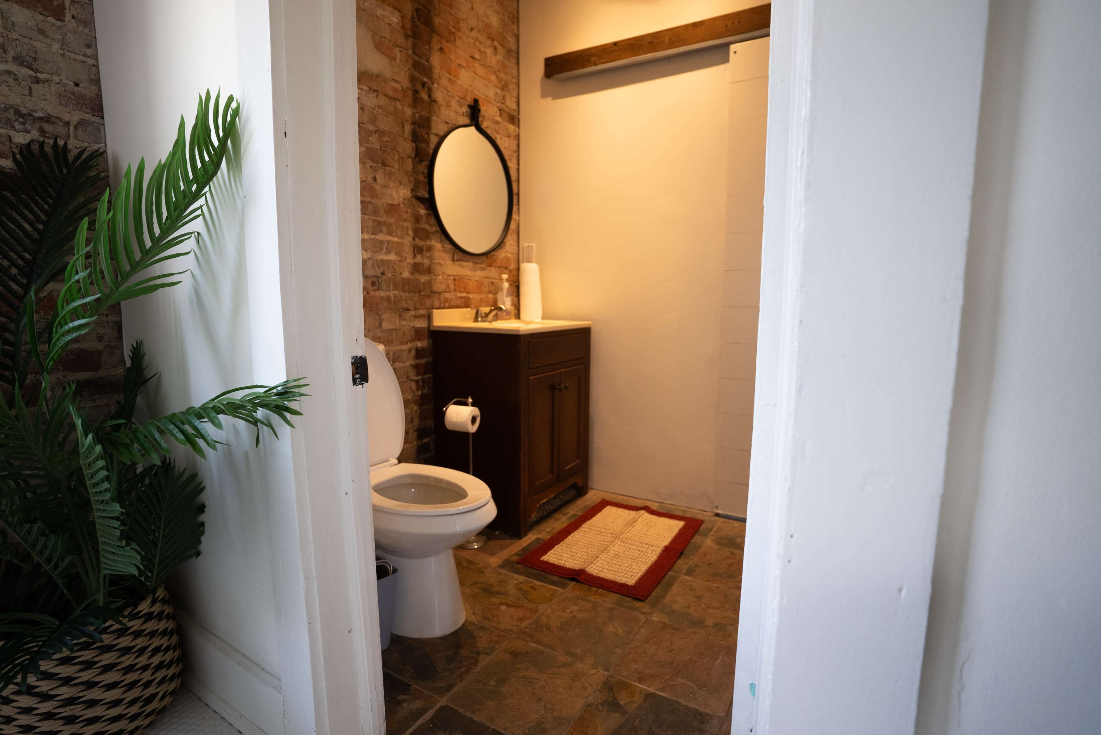 The image shows a small bathroom with a toilet, sink, and a mirror, featuring brick walls and a fern plant near the entrance.