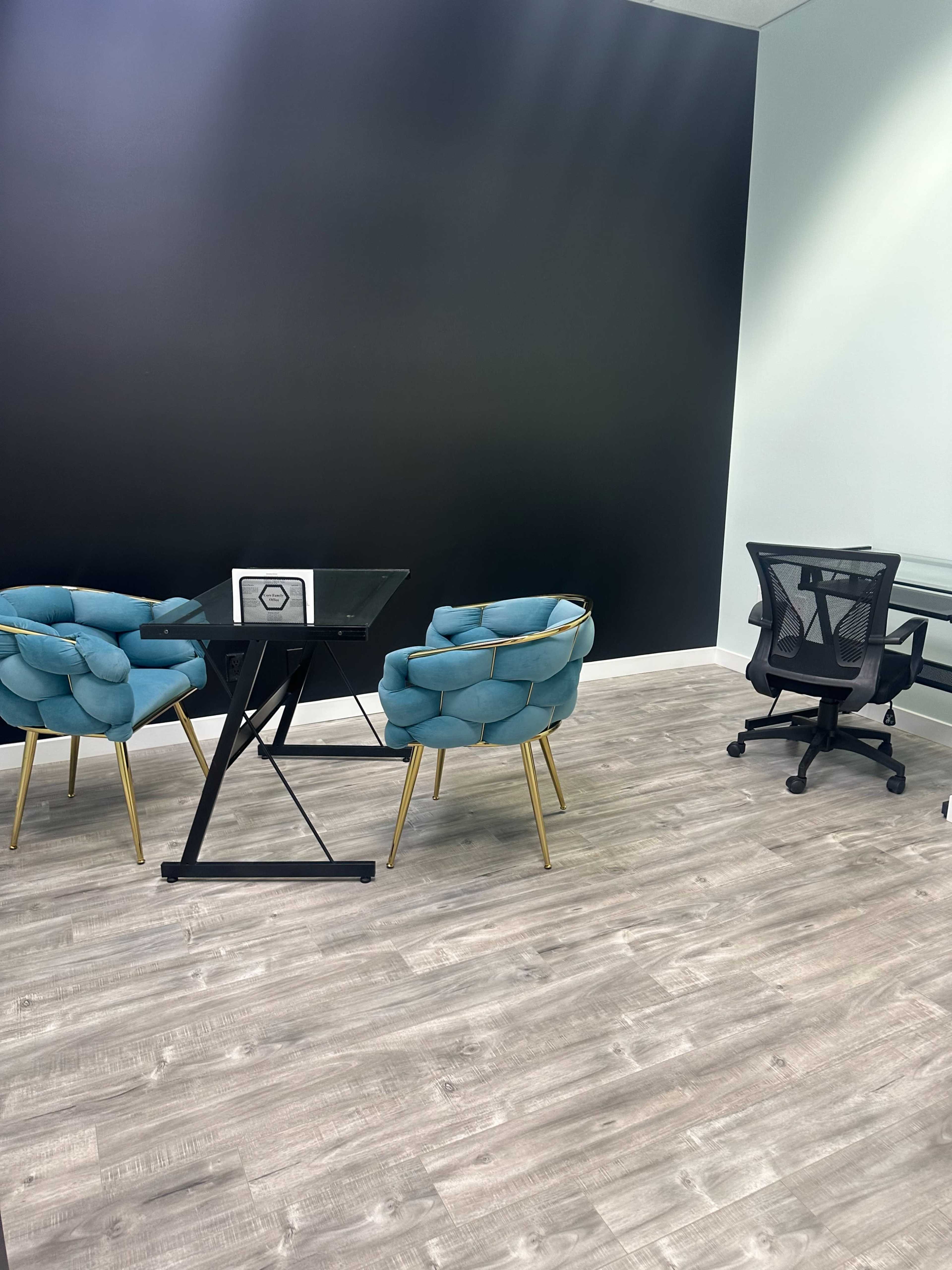 A modern office space with two blue chairs and a glass table, a black accent wall, and a black office chair positioned nearby.