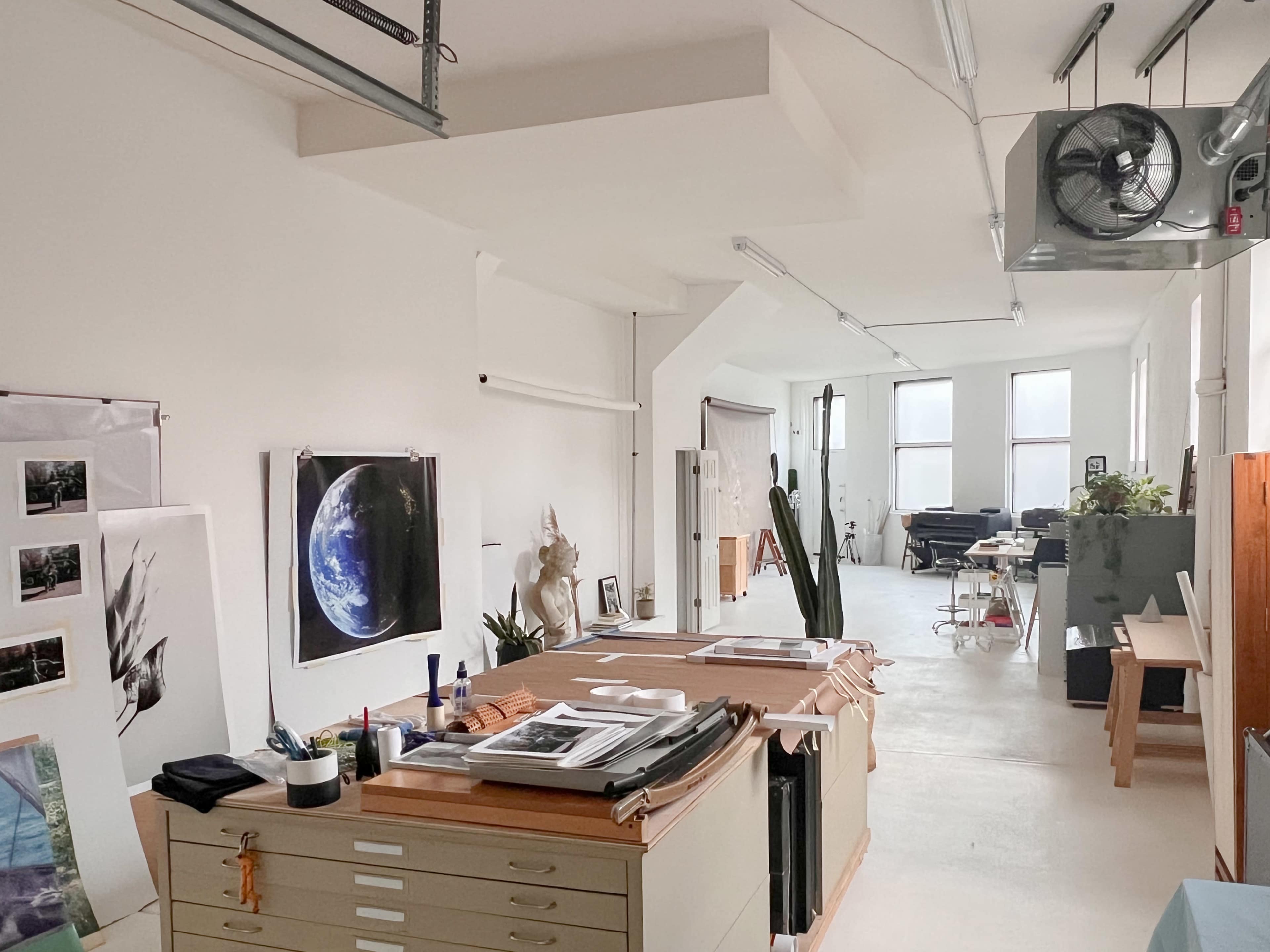 A bright art studio with several worktables, an assortment of artwork on the walls, and a view of additional creative spaces in the background.