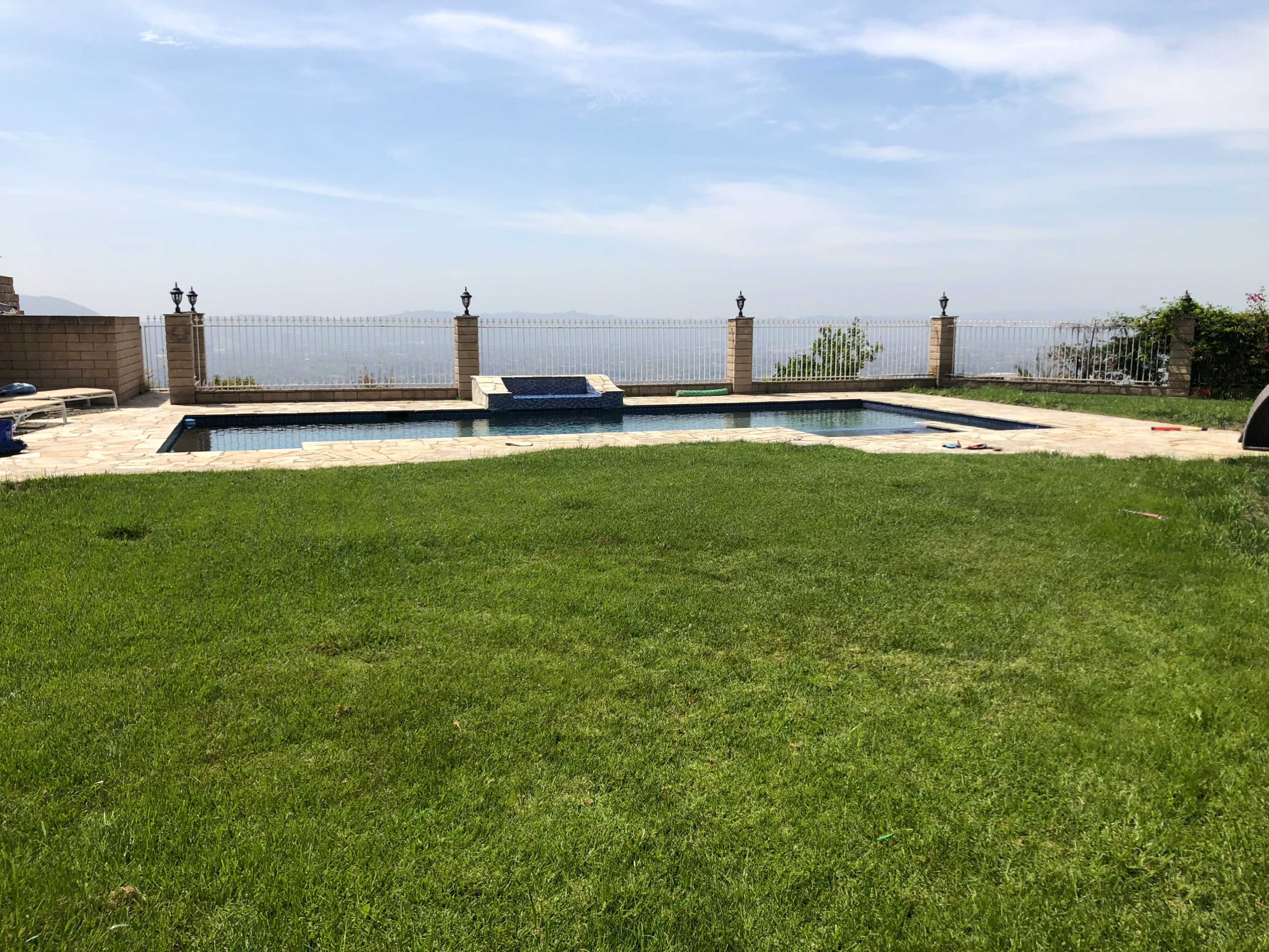 The image shows a rectangular swimming pool surrounded by a well-maintained grassy area and a fence overlooking a distant view.