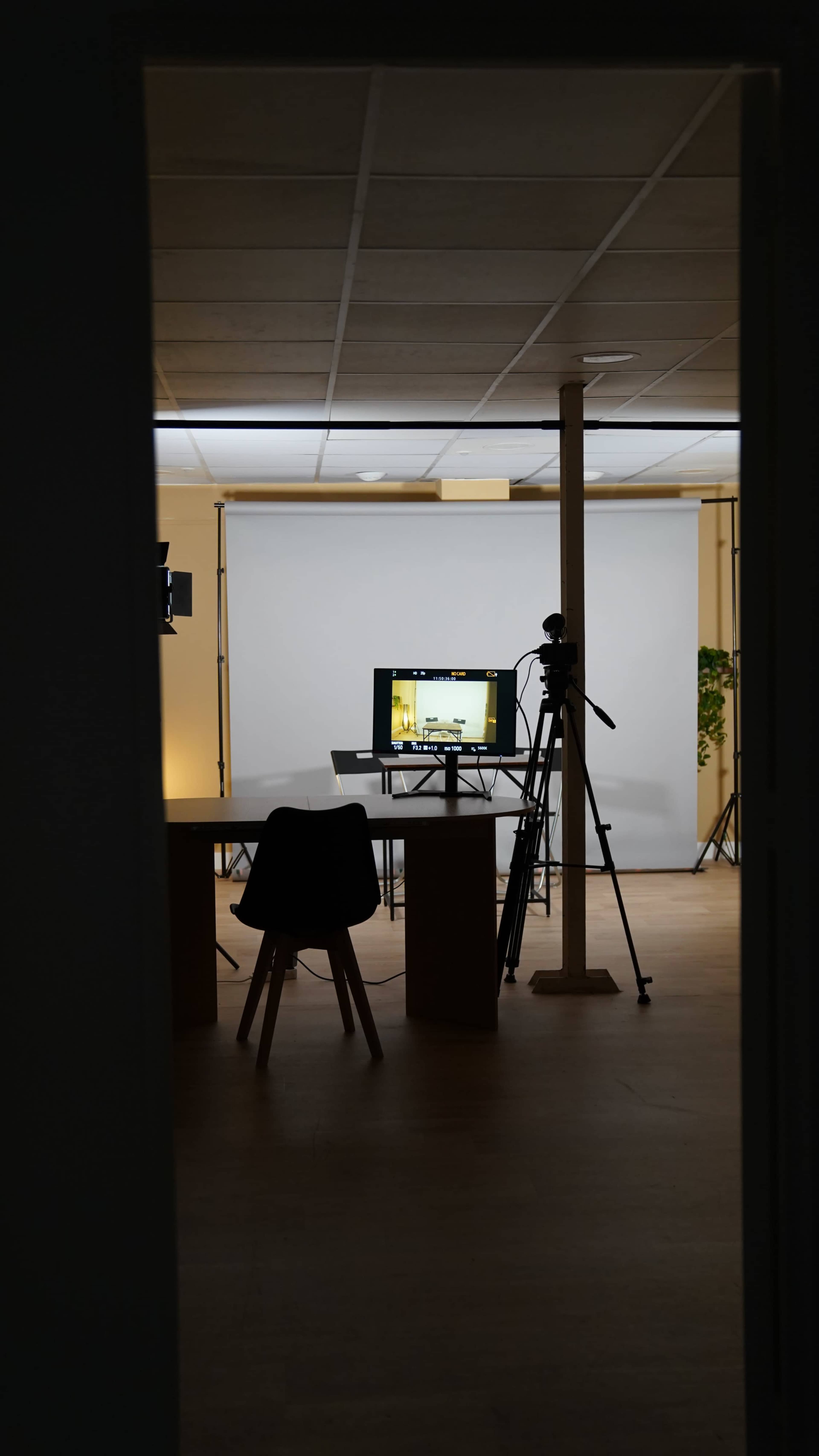 A dimly lit studio setup features a table and chair in the foreground, with a computer monitor and camera on a tripod visible in the background.