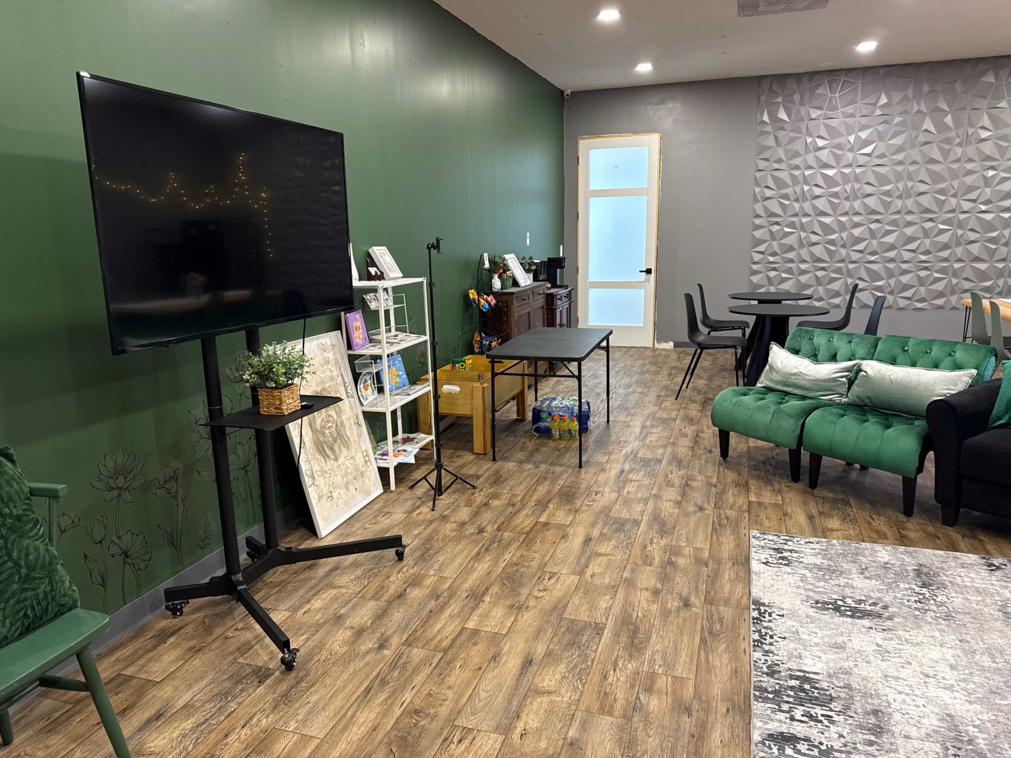 The image shows a modern indoor space with green walls, a large screen on a stand, a table, chairs, and a couch arranged for relaxation or socializing.