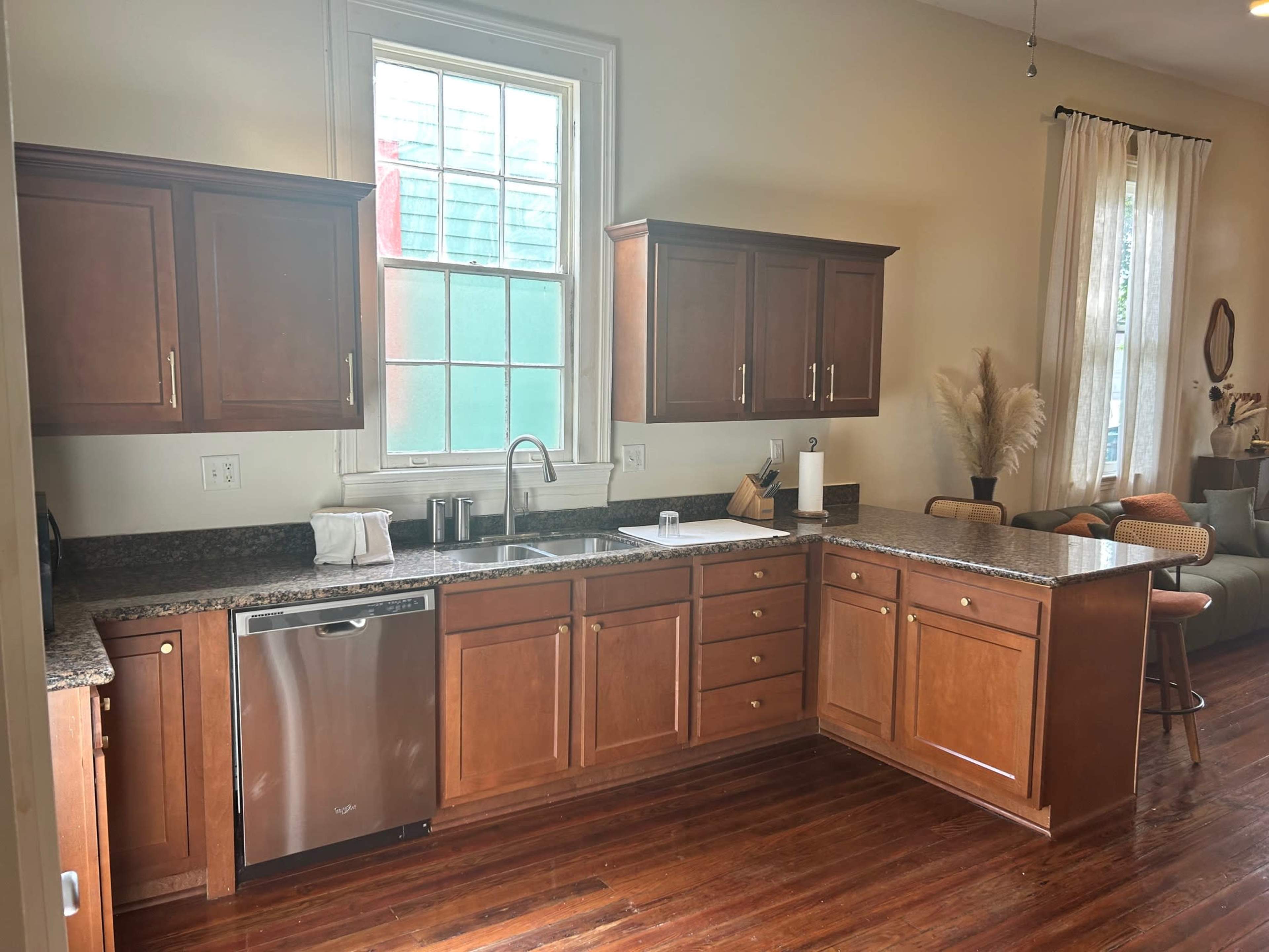 The image shows a tidy kitchen with wooden cabinets, a stainless steel dishwasher, and a granite countertop, featuring a sink and a window.
