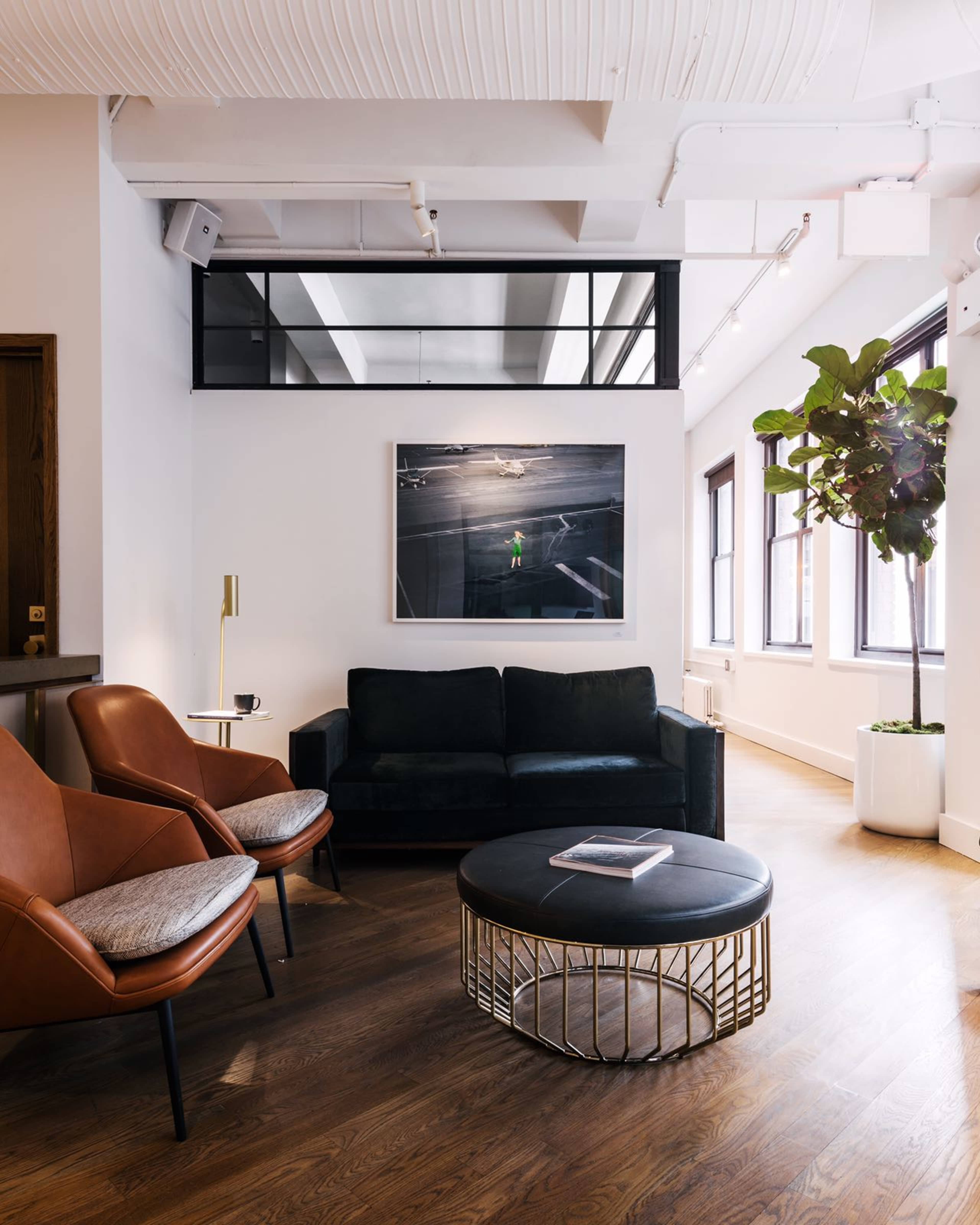 A modern lounge area featuring a black sofa, two brown accent chairs, a round coffee table with a gold base, and large windows allowing natural light.