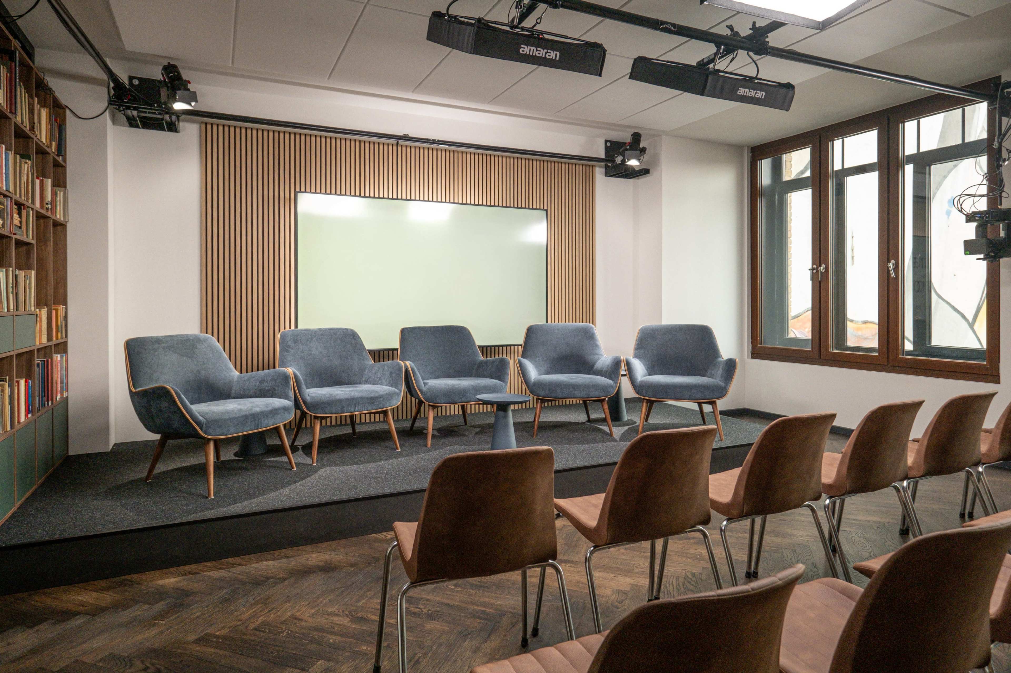A modern stage setup features six blue chairs arranged in a semi-circle on a dark platform, with a chalkboard and large windows in the background.