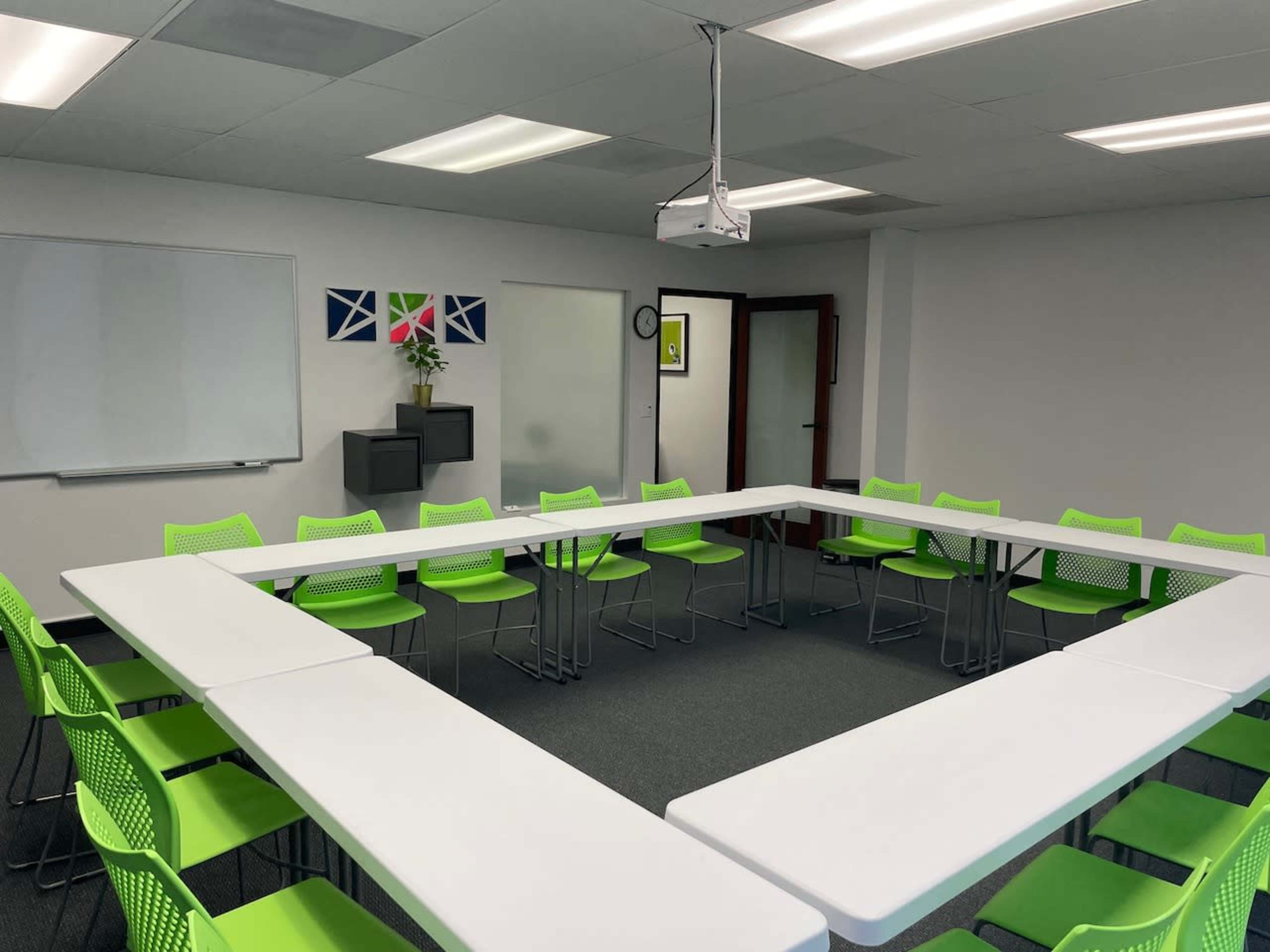 A conference room features a white table arranged in a square shape with bright green chairs around it, along with a projector and whiteboard on the walls.