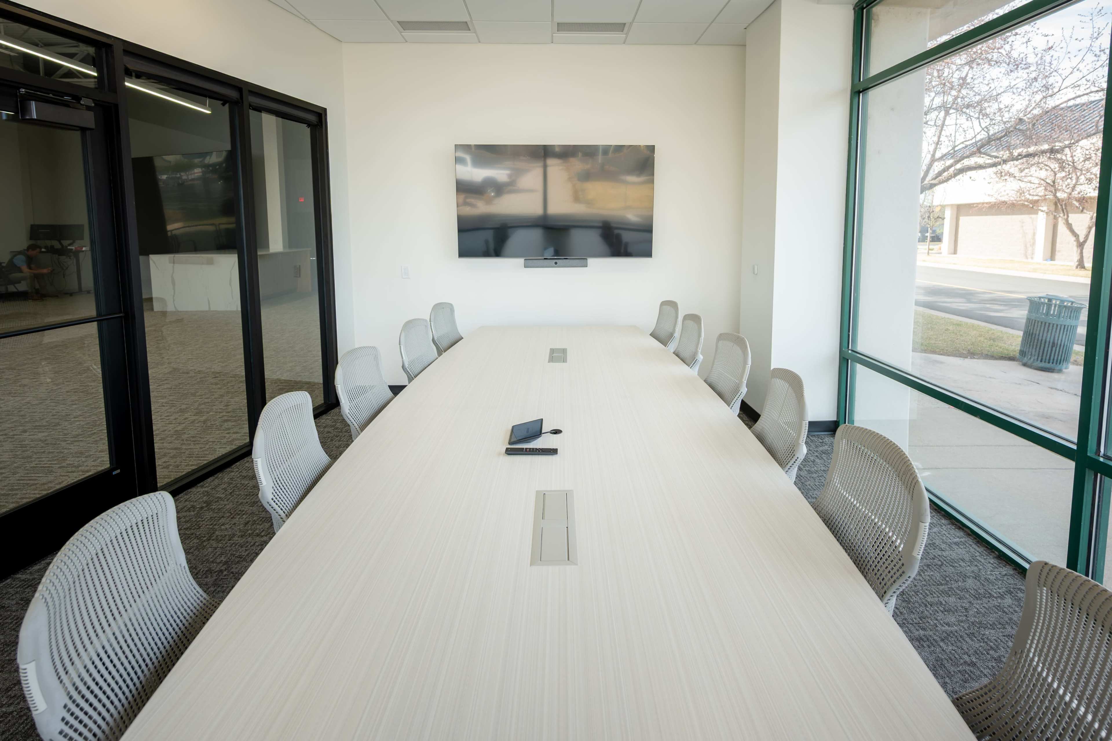A modern conference room features a long table with ergonomic chairs and a wall-mounted television.