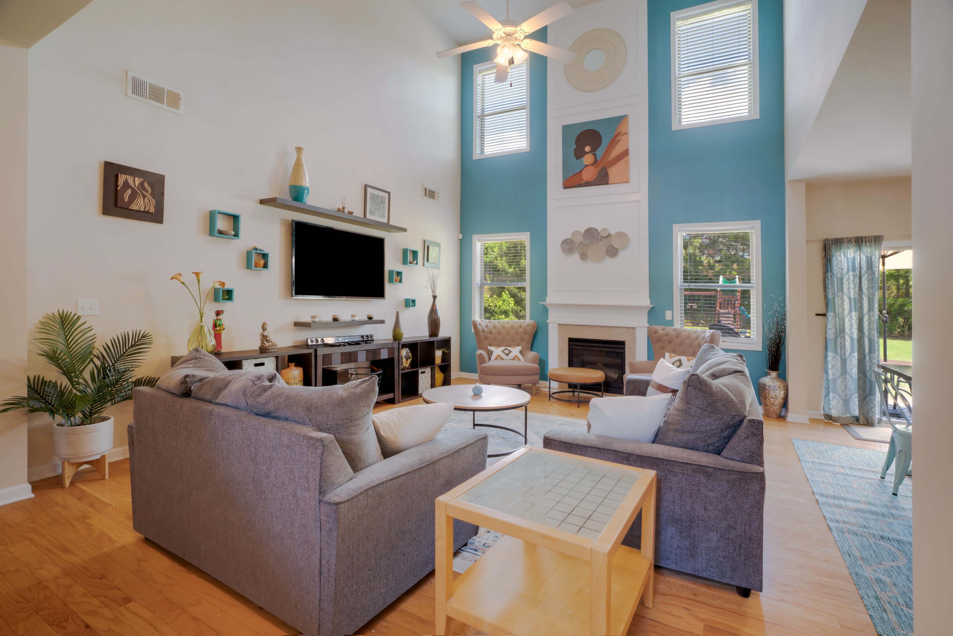 The living room features a central seating area with gray sofas, a wooden coffee table, and a decorative TV unit against a blue wall, accented by large windows and artwork.
