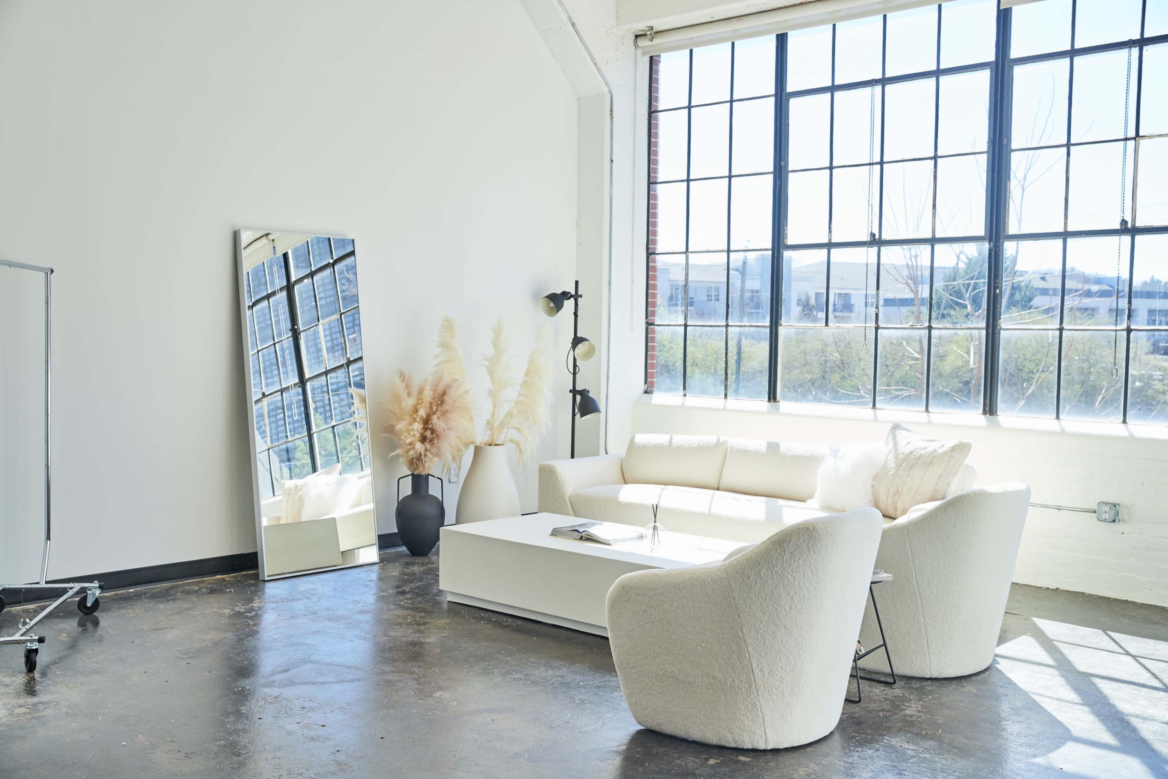 A bright, minimalist living space with a white sofa, a large mirror, a coffee table, and decorative plants, illuminated by natural light from large windows.