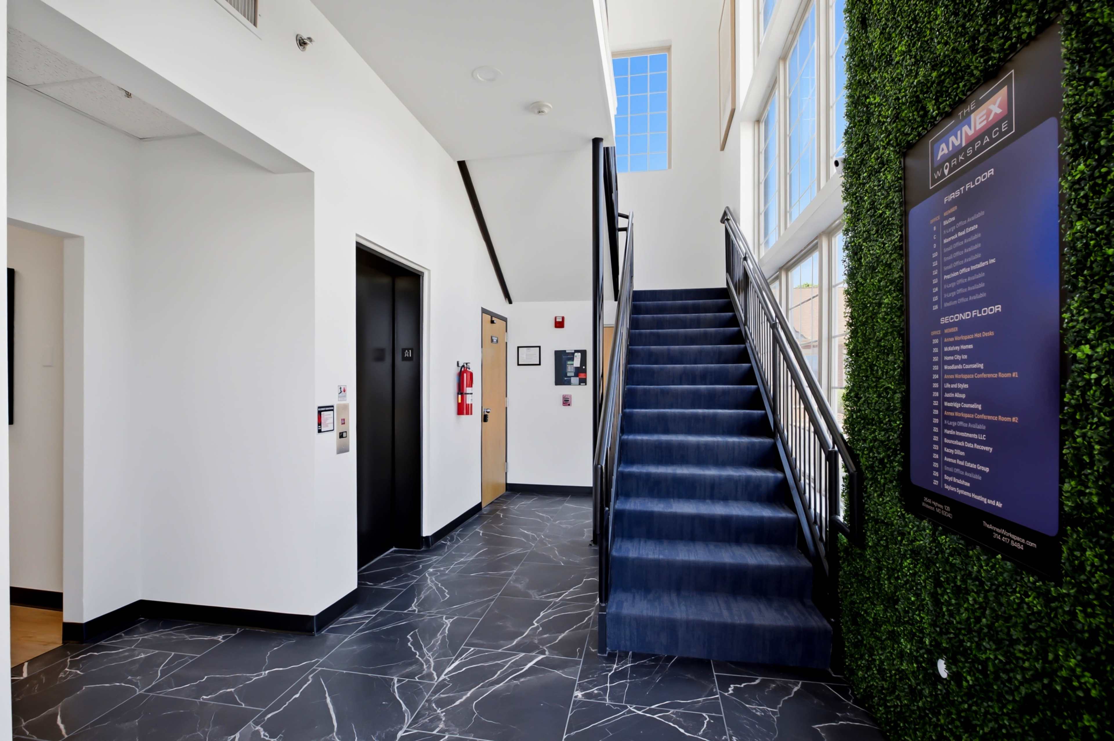 The image shows a modern interior lobby with an elevator, a staircase leading to an upper floor, and a decorative green wall.