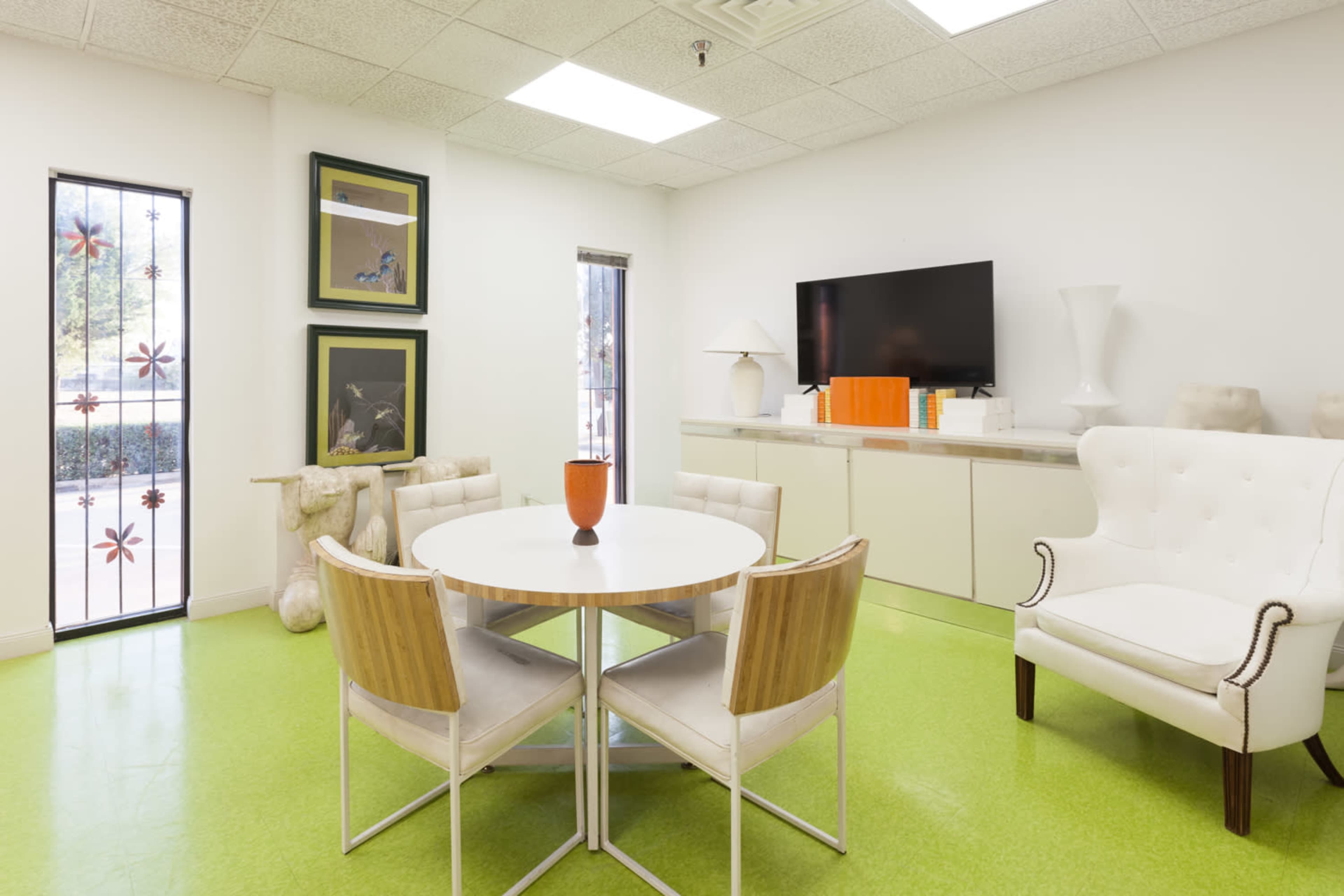 A modern office space features a round table with four chairs, a large television mounted on the wall, and decorative art pieces arranged on shelves.