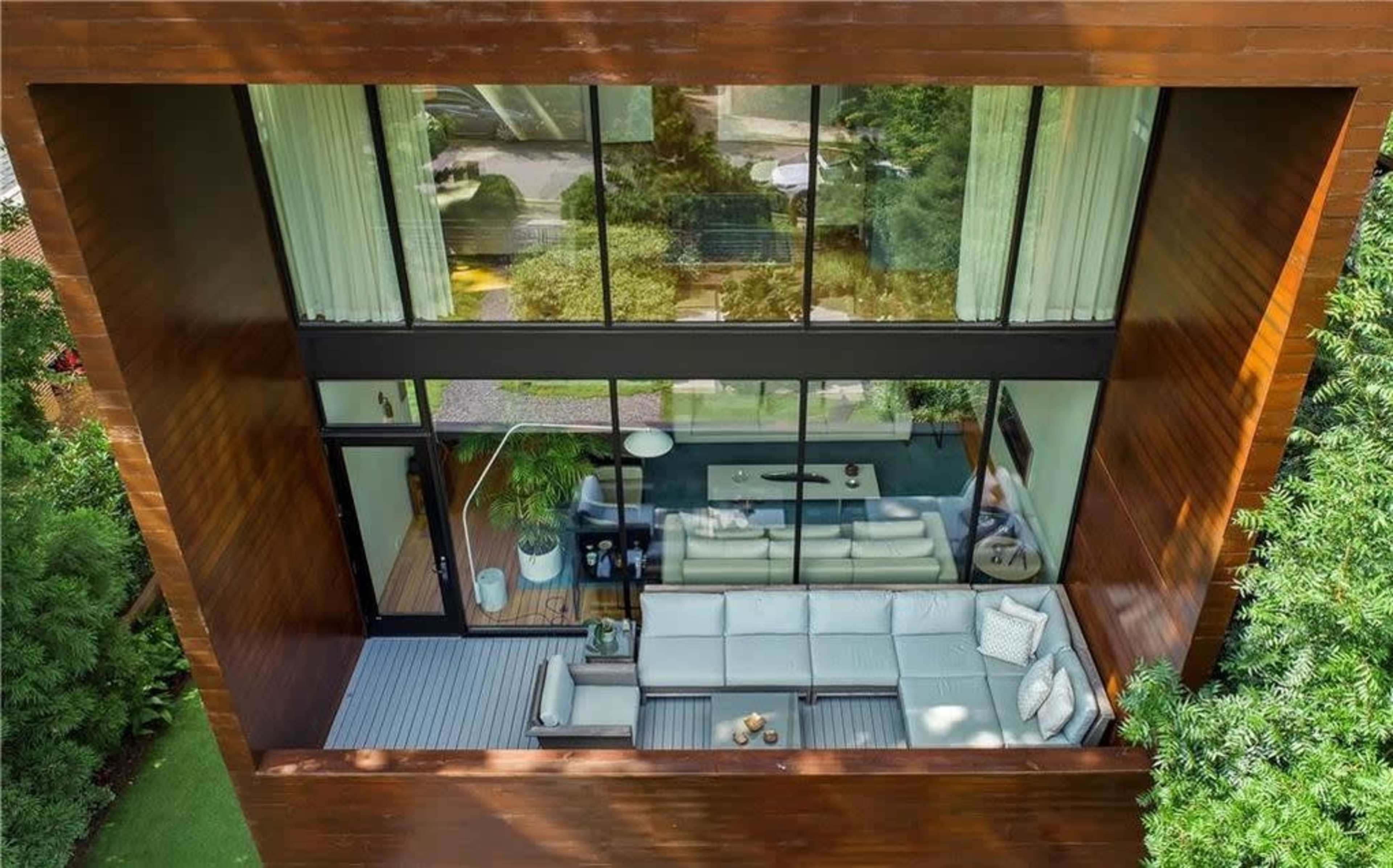 The image shows a modern, open-concept living space with large glass windows and a wooden exterior, furnished with a light-colored sectional sofa.