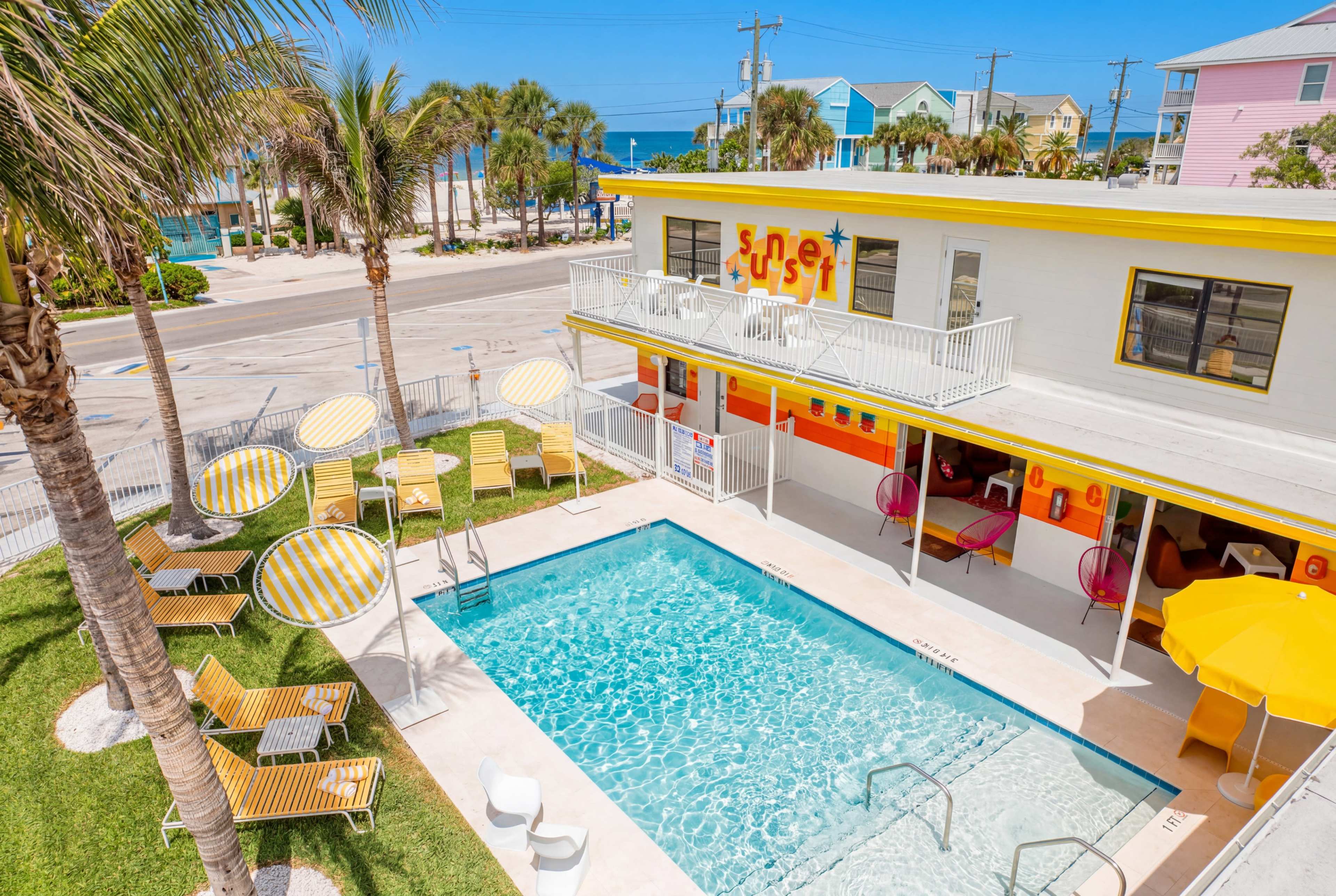 The image shows a colorful pool area at a motel, featuring a clear blue swimming pool surrounded by lounging chairs and umbrellas, with the ocean visible in the background.
