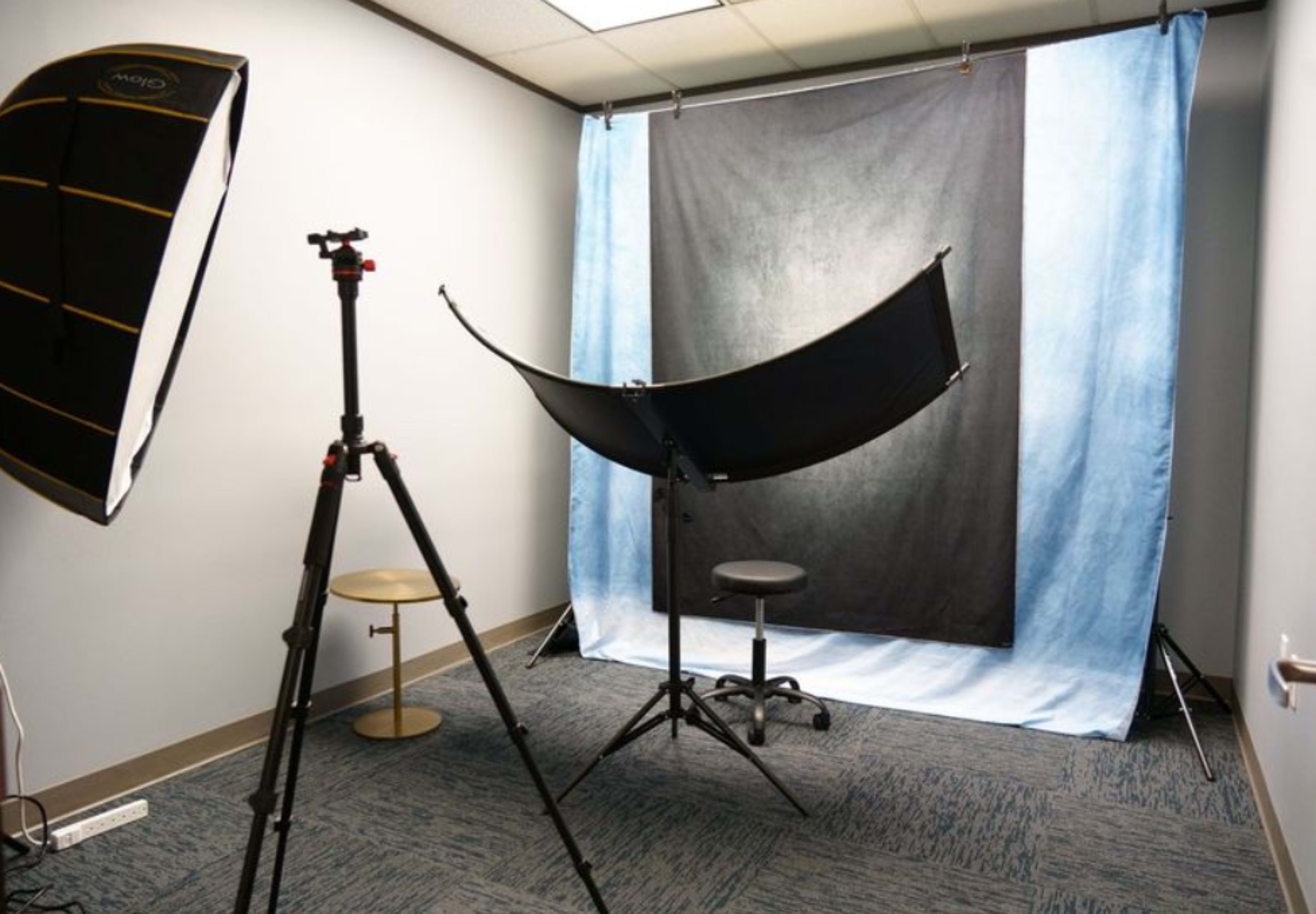 The image shows a photography setup in a small room, featuring a backdrop with blue and black fabric, a tripod with a light modifier, and a stool positioned for a subject.