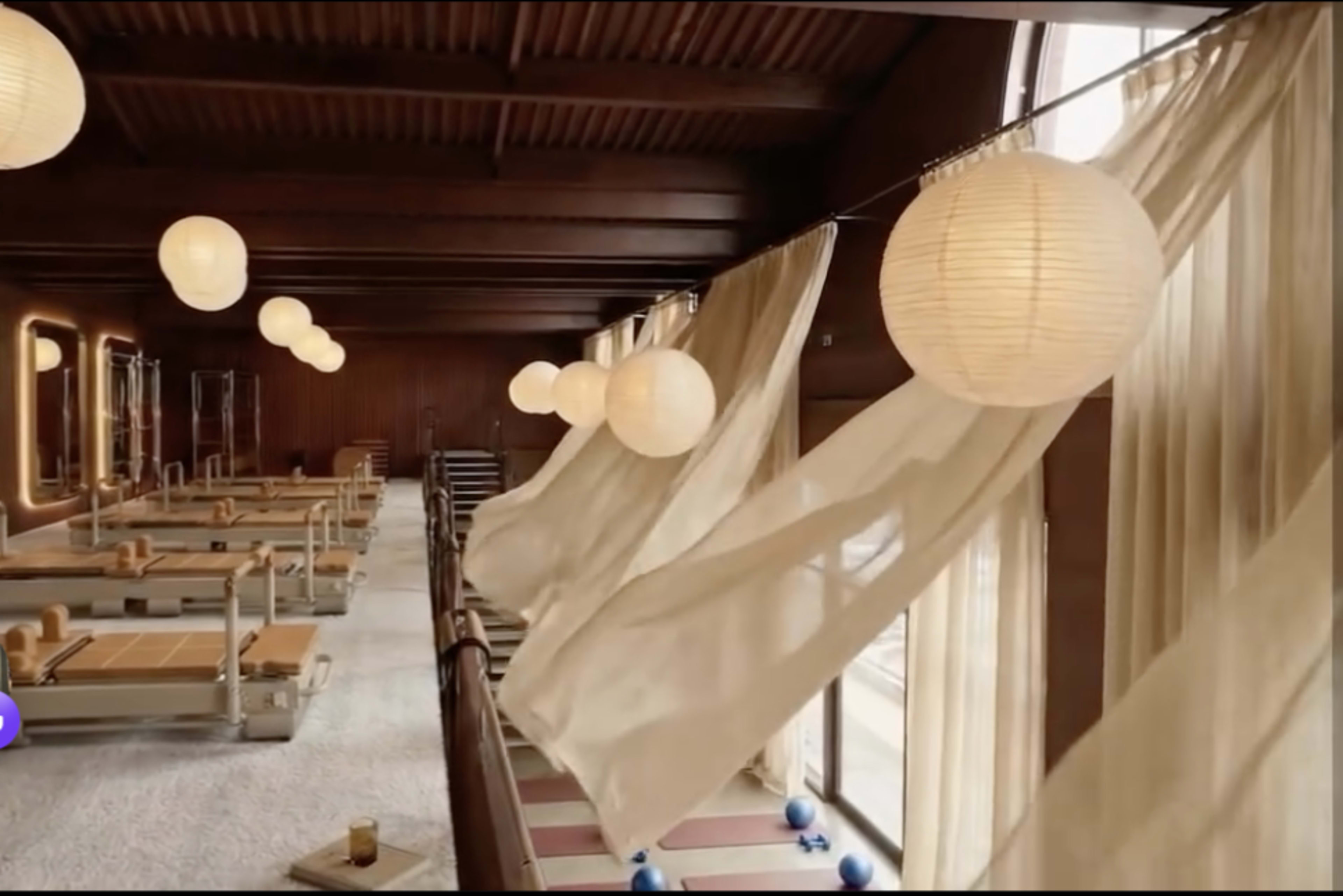 The image shows a spacious, wooden-floored fitness studio with reformer machines, large windows, and sheer curtains blowing in the breeze.