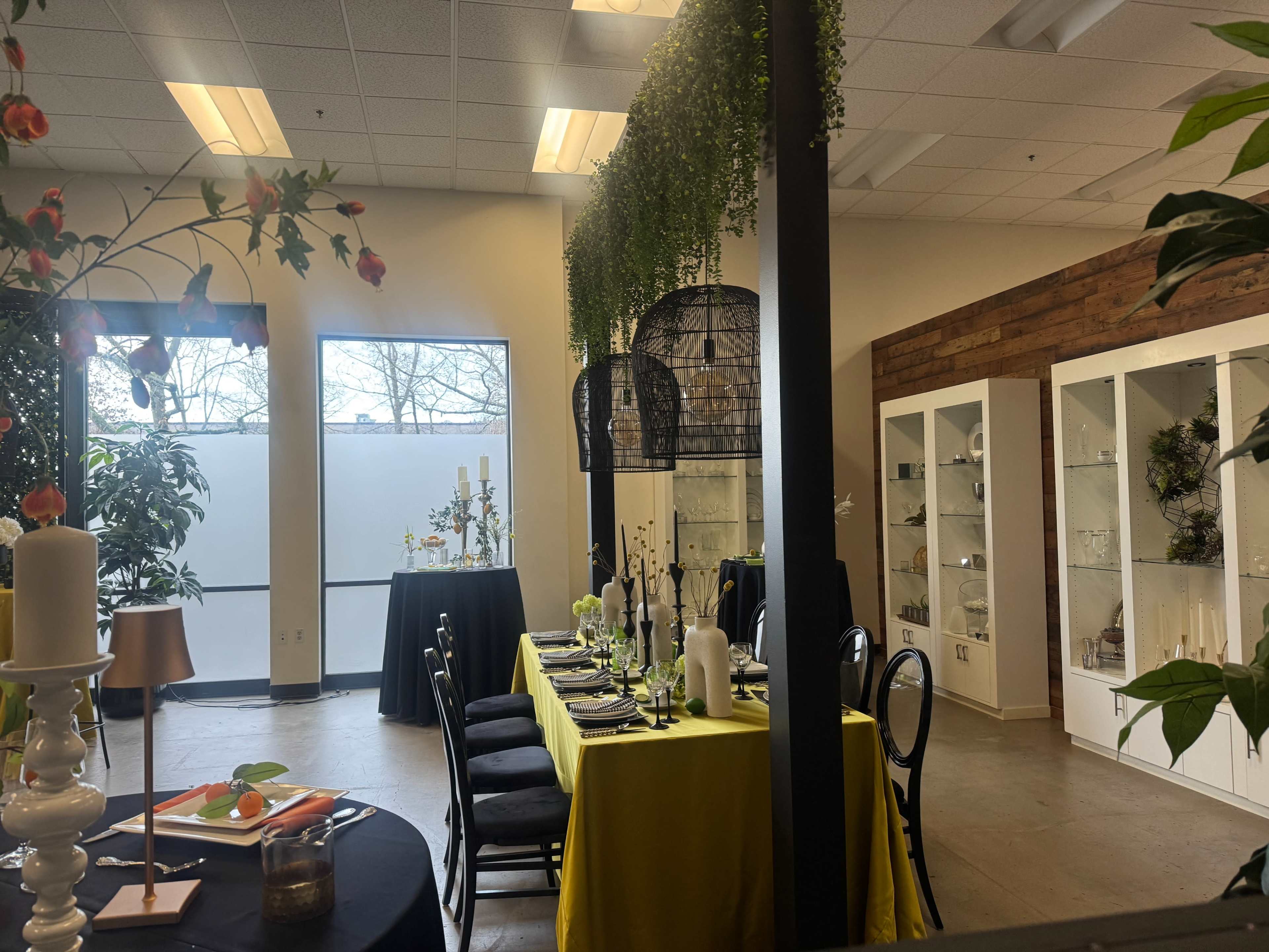 A brightly lit event space features two elegantly set dining tables surrounded by decorative plants and display cabinets.