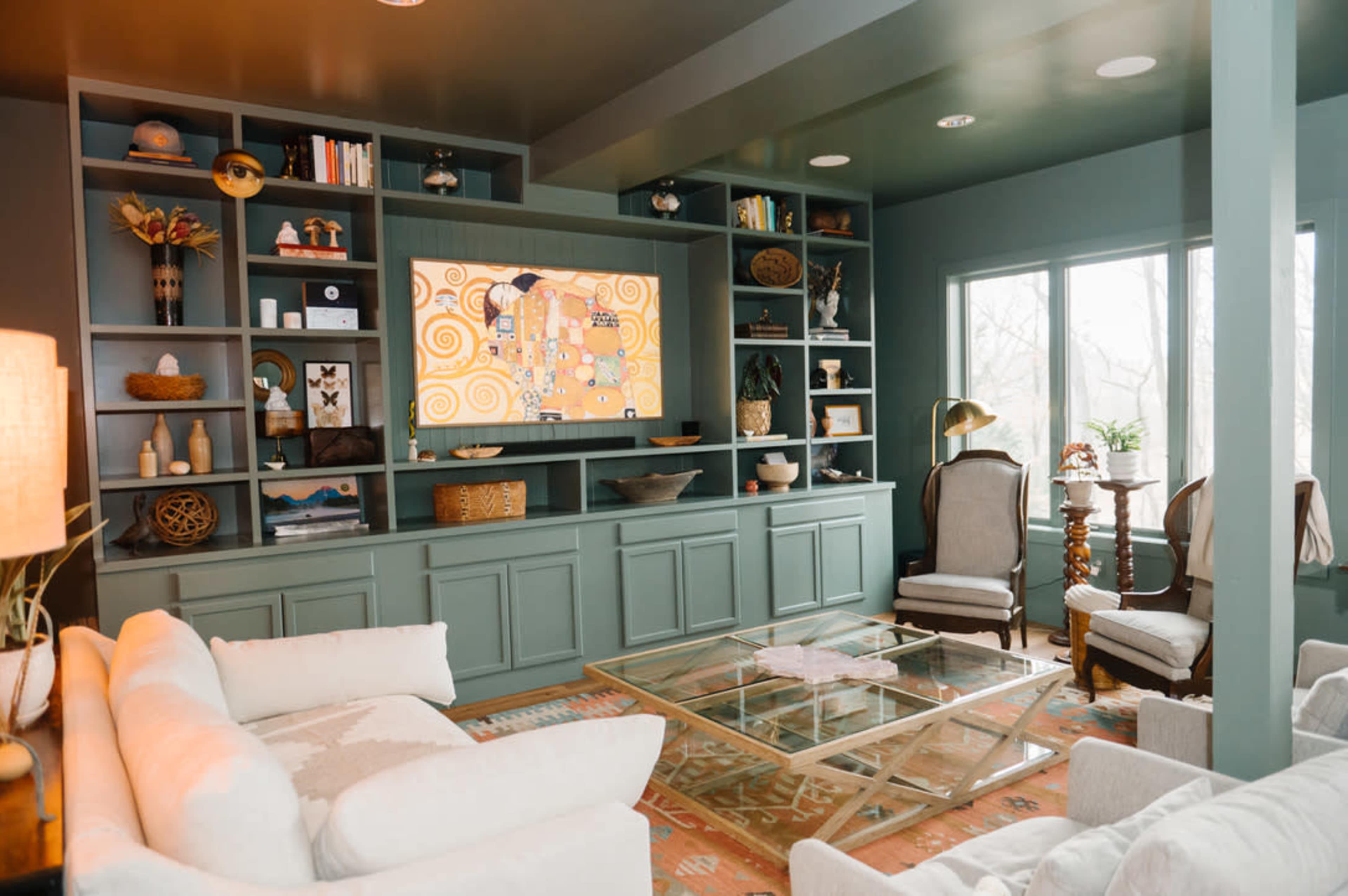 A modern living room features a large green built-in shelving unit with a television, a glass coffee table, and comfortable seating arranged near large windows.