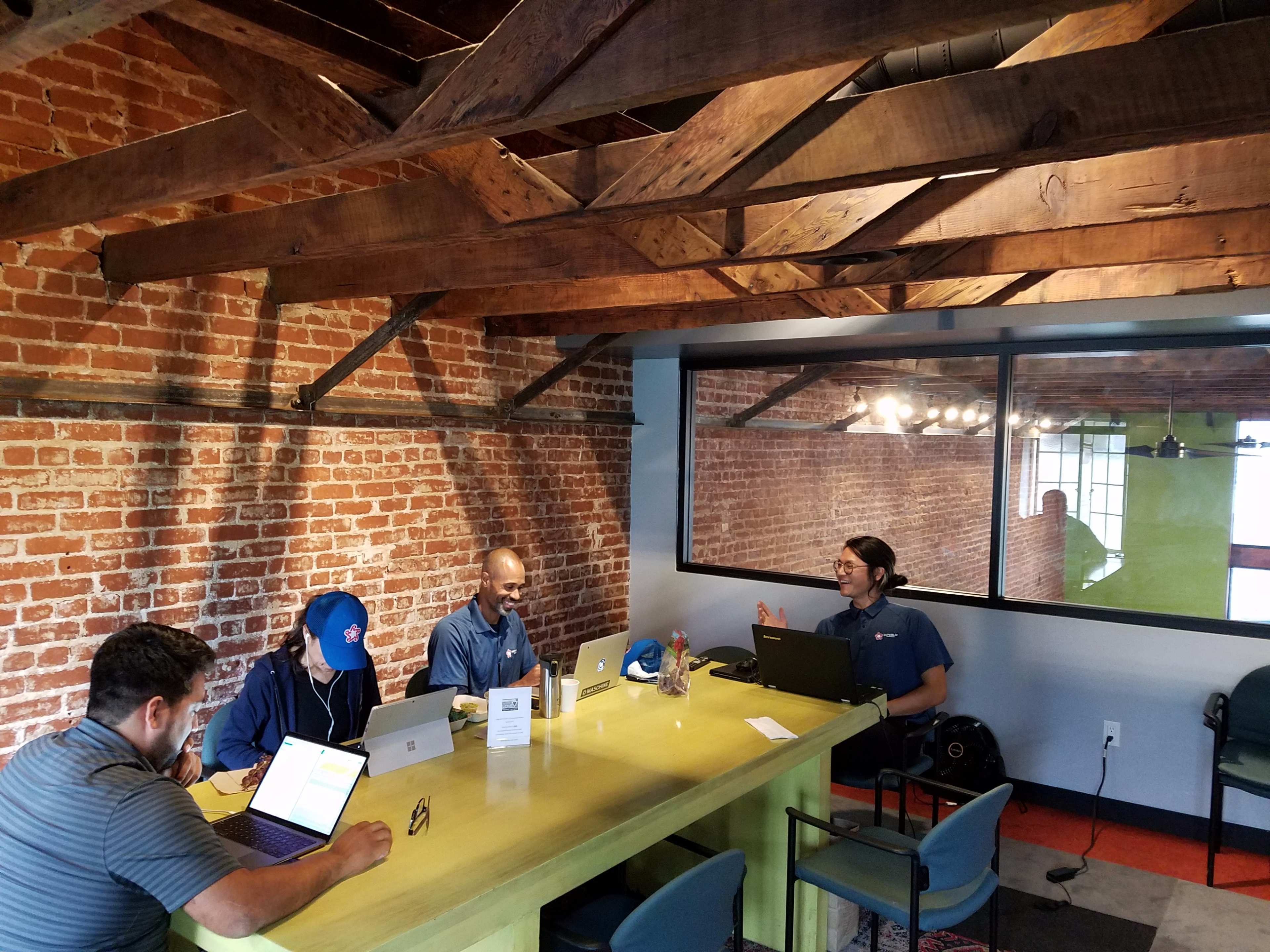 A group of four people work on laptops around a green table in a room with exposed brick walls and wooden beams.