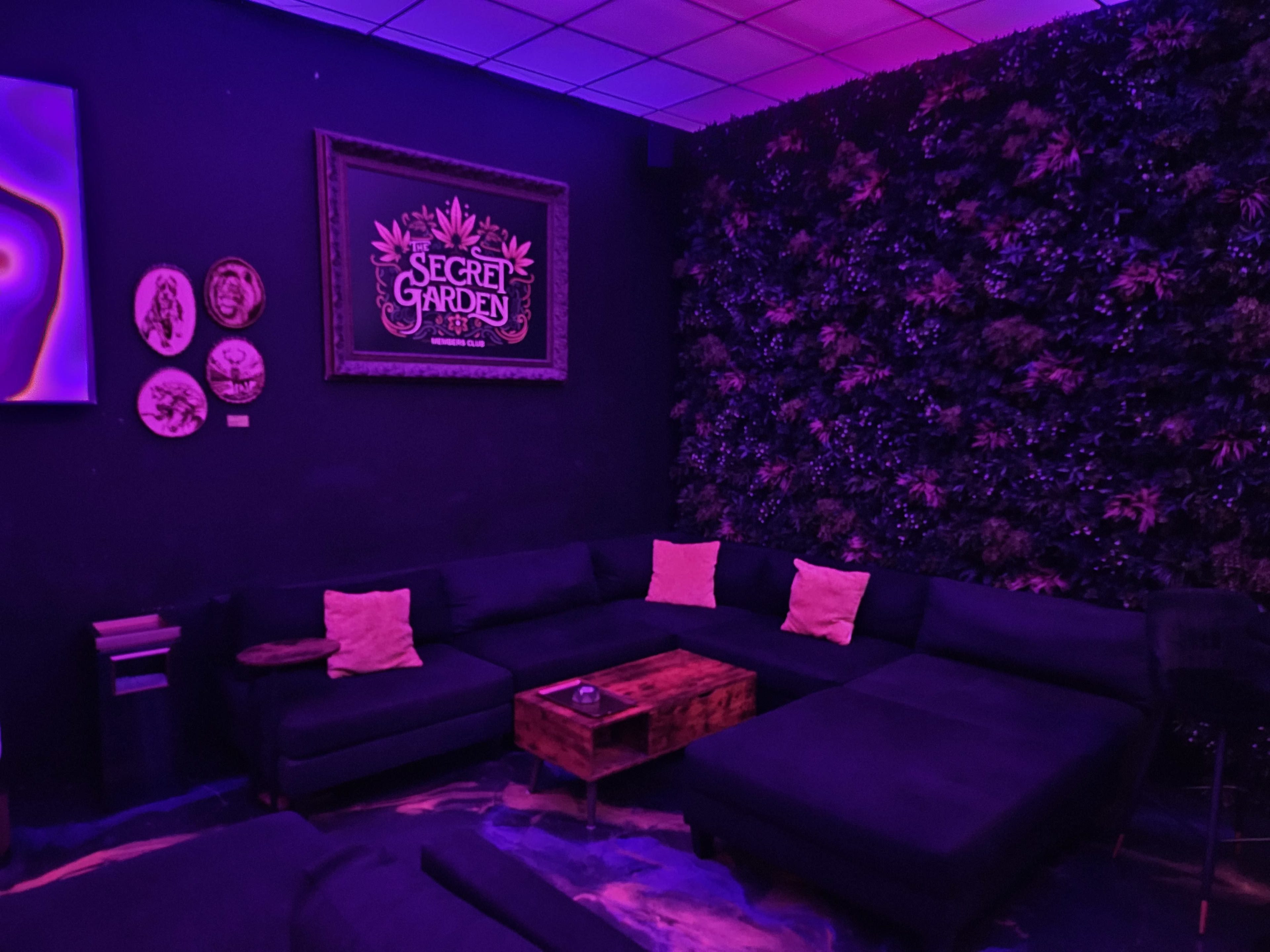 The image shows a lounge area with dark seating, a wooden coffee table, and a wall adorned with artificial greenery, illuminated by purple lighting.