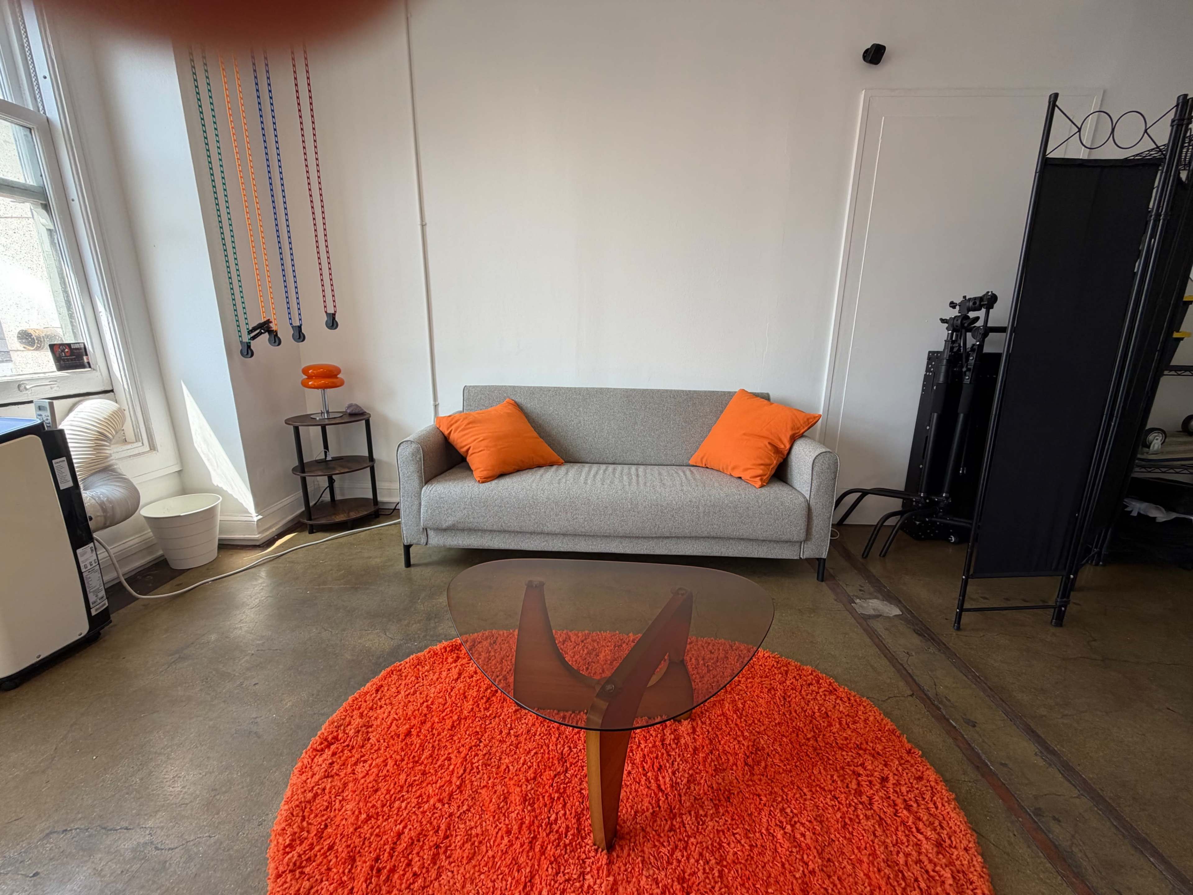 The image depicts a simple indoor space featuring a gray sofa with orange cushions, an orange circular rug, and a glass coffee table.