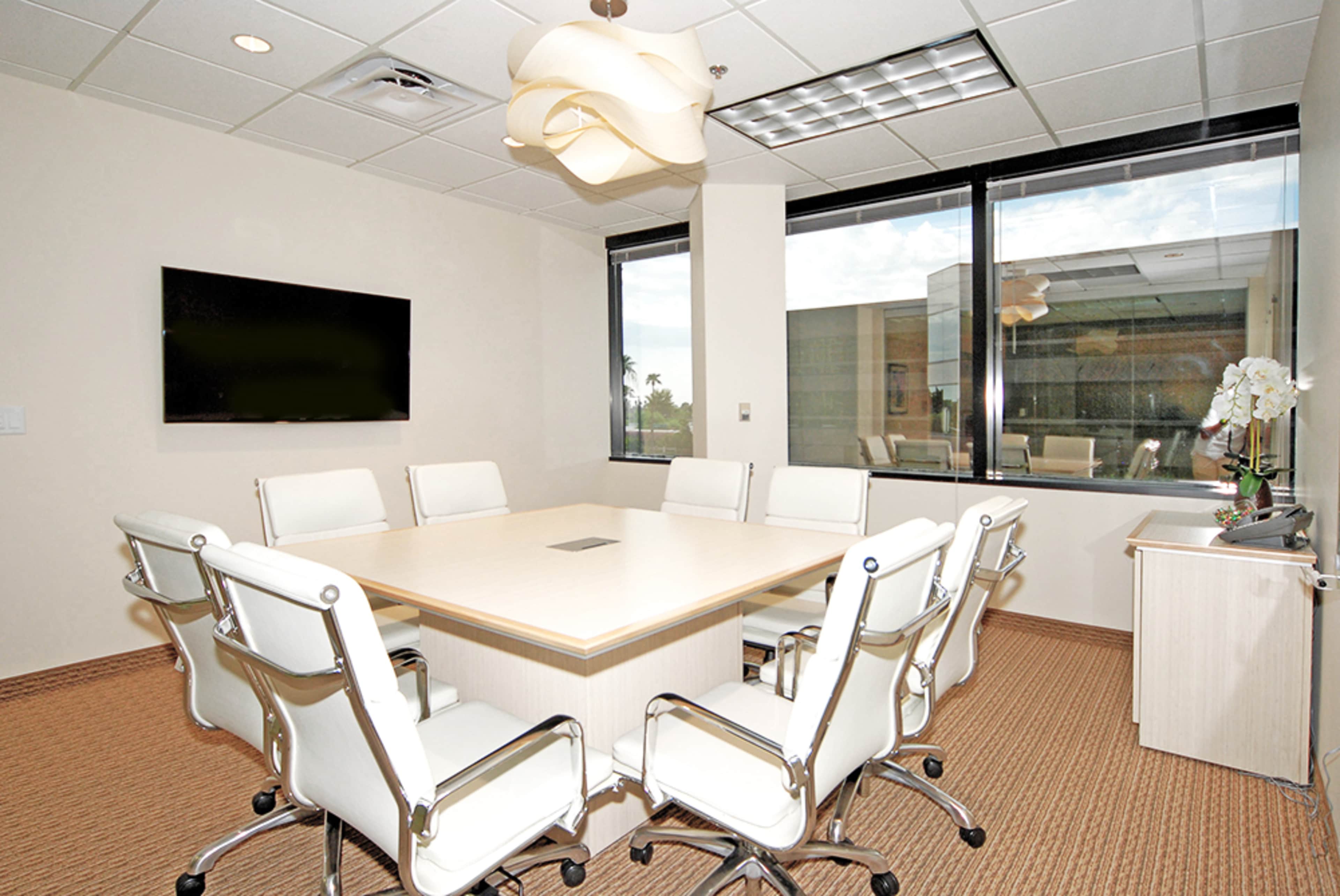 A modern conference room features a large wooden table surrounded by white chairs, with a television mounted on one wall and large windows letting in natural light.