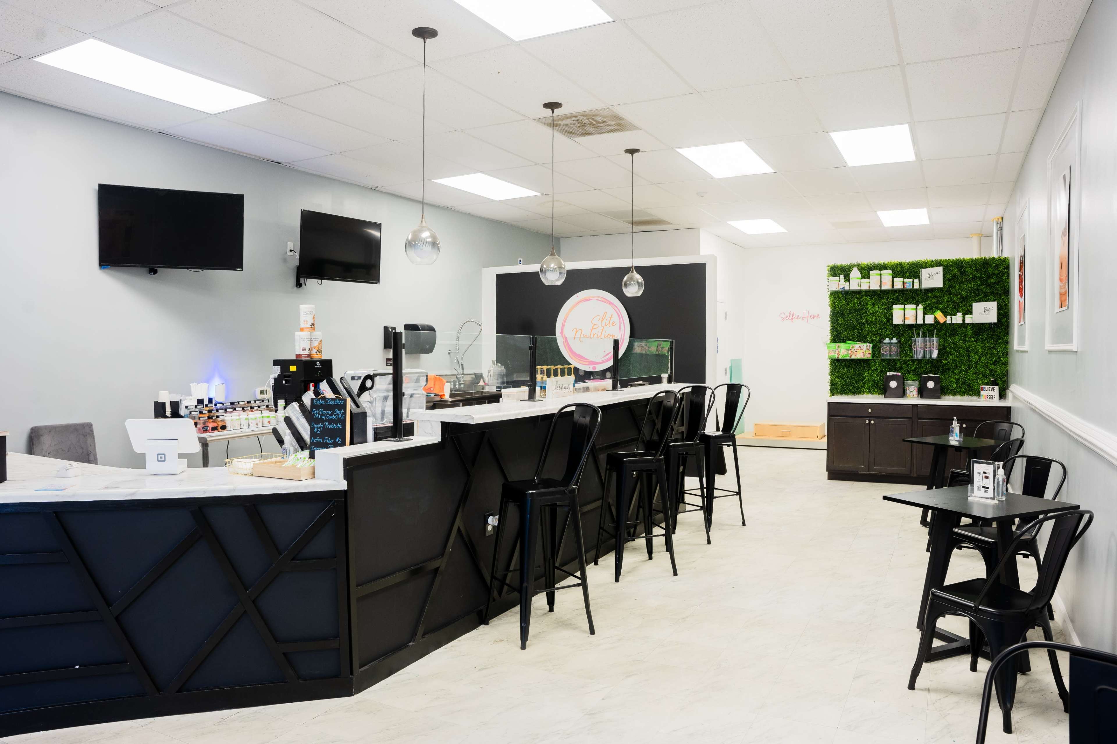 The image shows a modern café interior with a dark counter, several bar stools, green wall decor, and two televisions mounted on the wall.